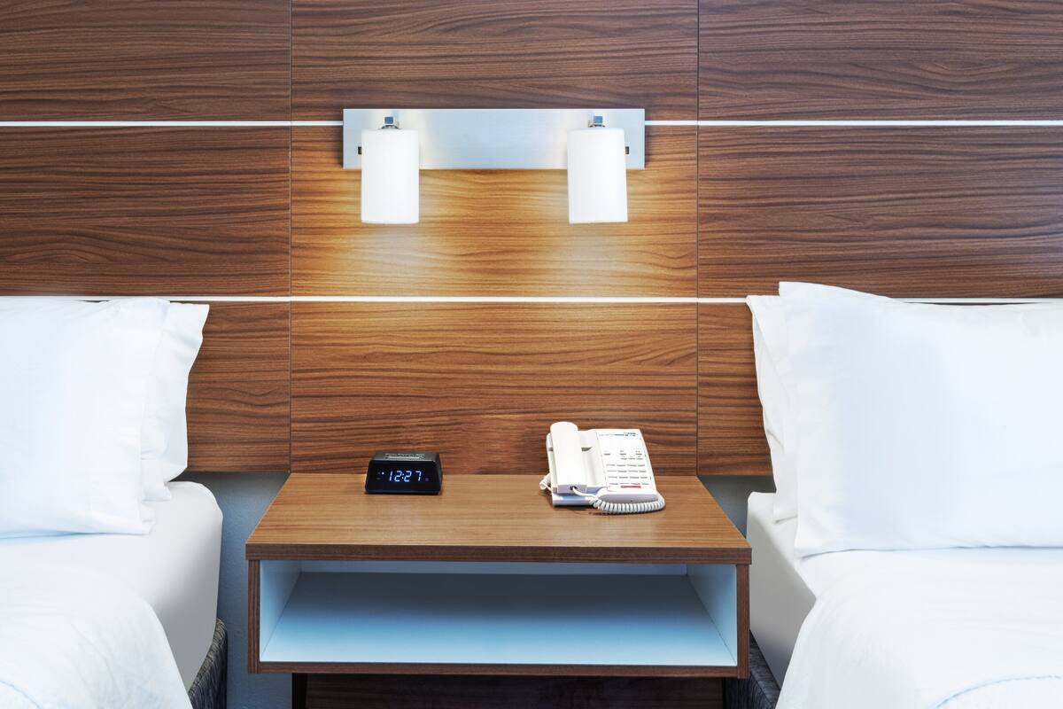 Une chambre d’hôtel avec une tête de lit en bois, deux tables de chevet, une horloge, un téléphone et du linge de lit blanc.