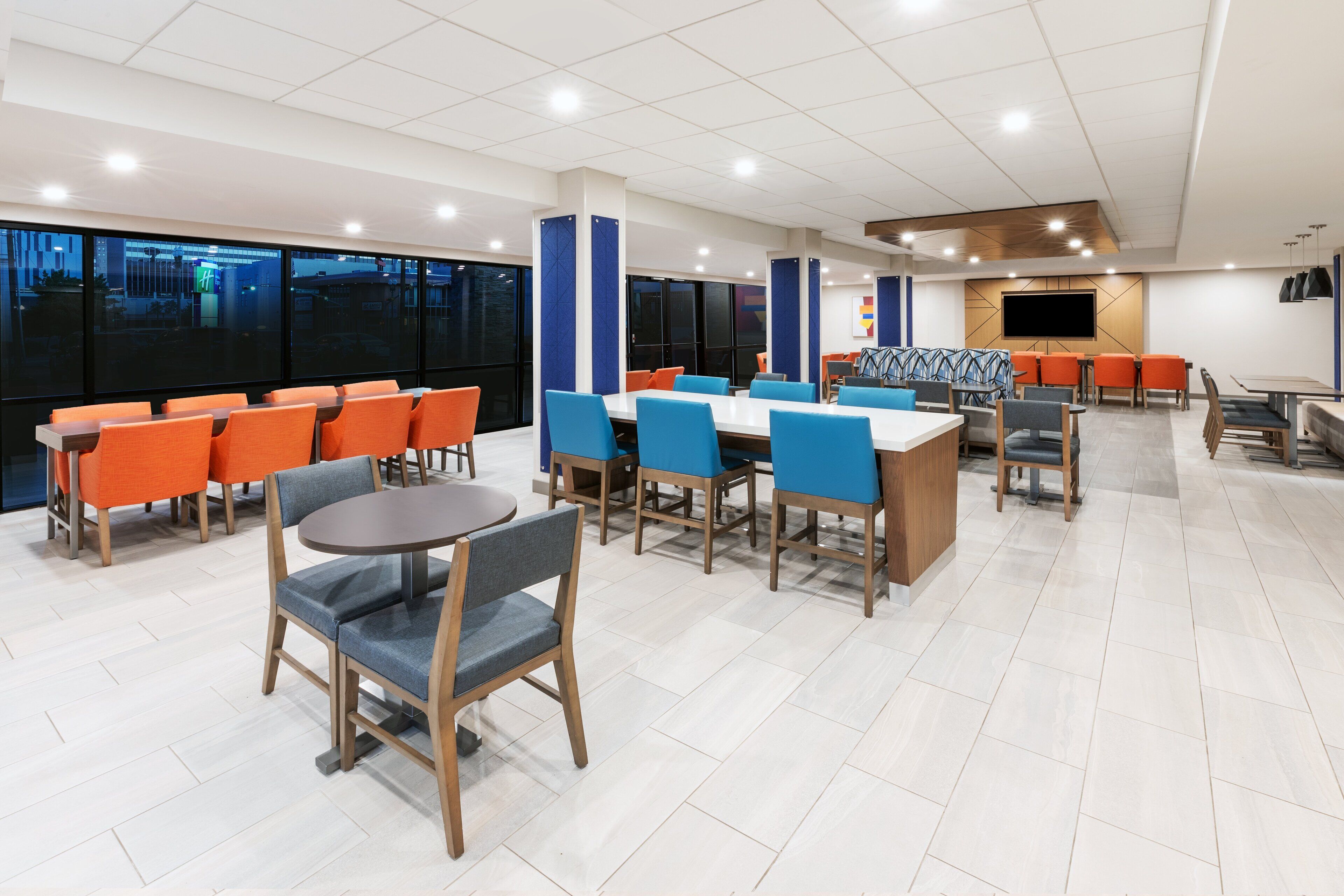 A modern dining area with multiple tables and chairs, large windows, and a flat-screen TV on the wall.