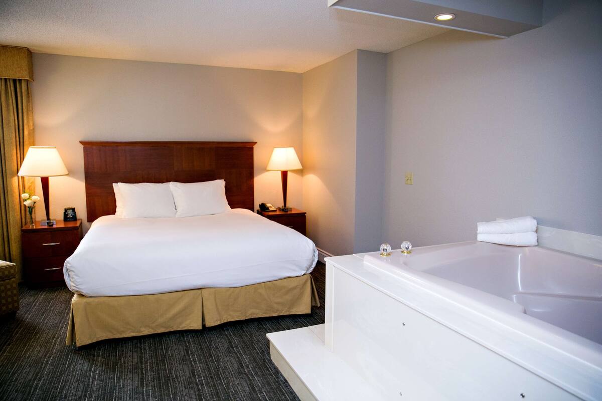 Habitación de hotel con una cama grande, dos lámparas de noche, una mesita de noche, un baño con bañera y piso alfombrado.