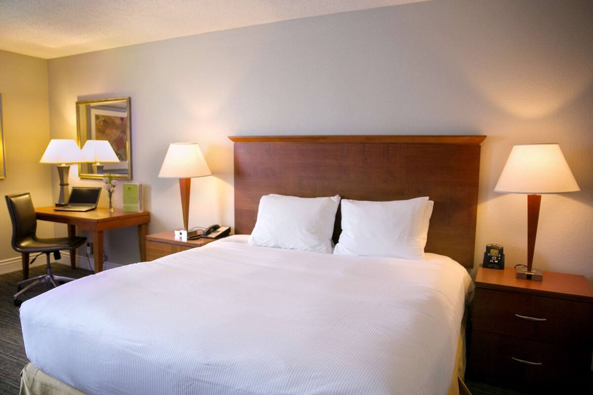 Habitación de hotel con una cama grande, dos lámparas de noche, un escritorio con una computadora portátil, una silla y una mesita de noche con un teléfono.