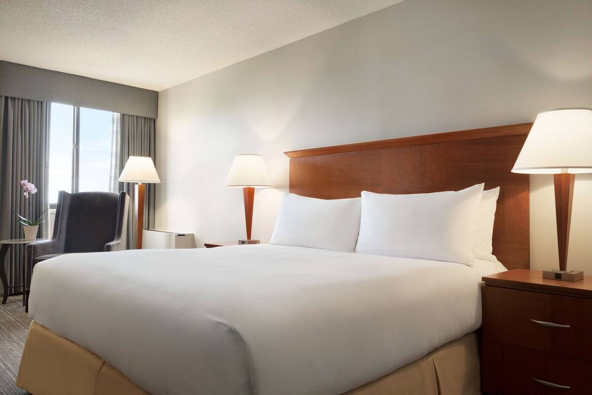 Habitación de hotel con una cama grande, dos lámparas de noche, cabecera de madera, un escritorio con una silla y una ventana con cortinas.