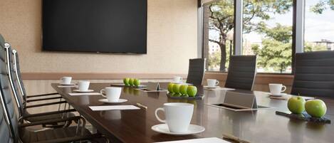 A conference room with a large table, chairs, a laptop, a projector screen, and a view of trees through the windows.