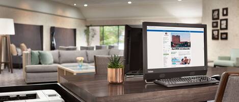 A modern hotel room with a desk, laptop, printer, and a view of the cityscape through large windows.