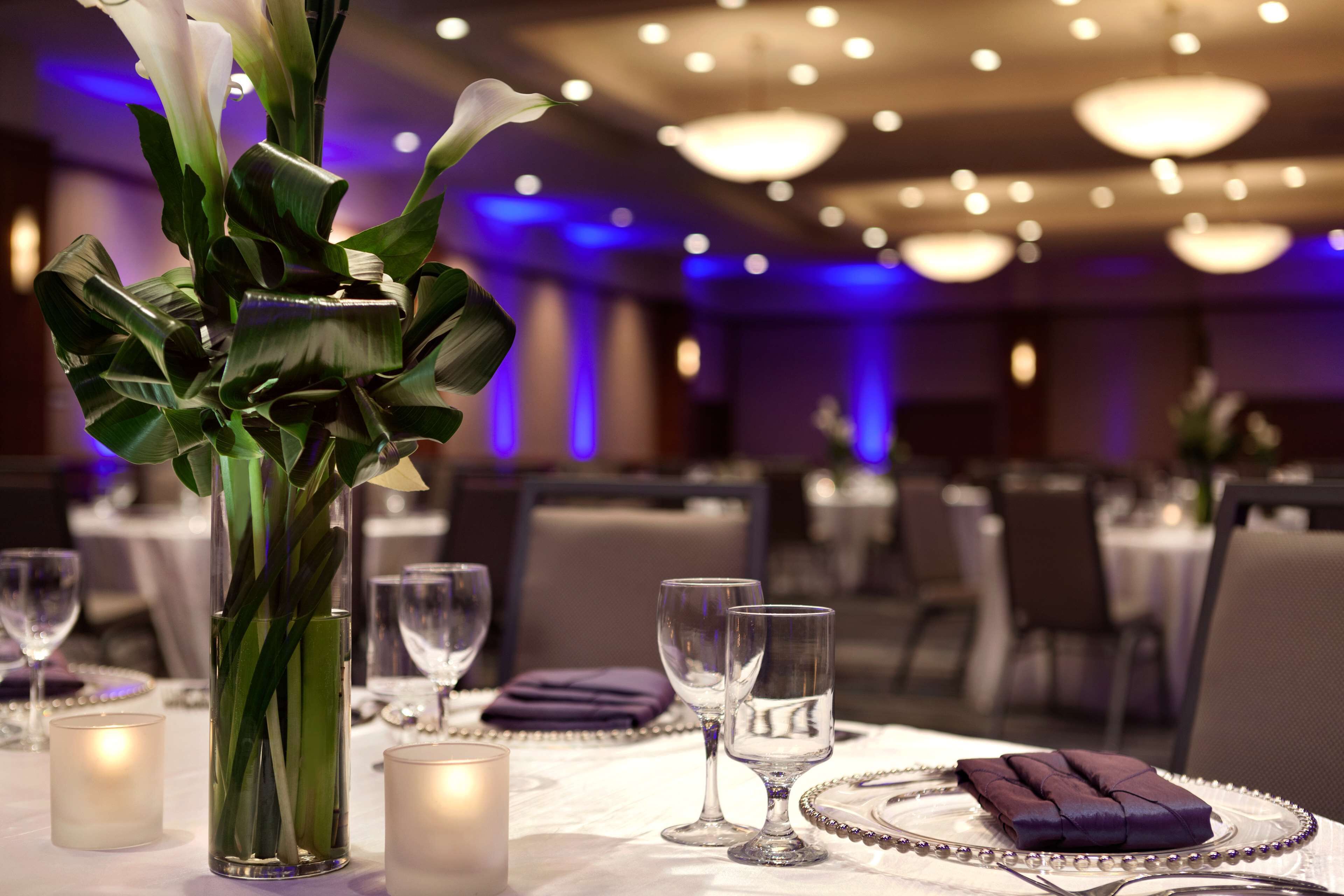 A dining area with tables set for two, featuring clear glassware and a centerpiece of white flowers in a glass vase.