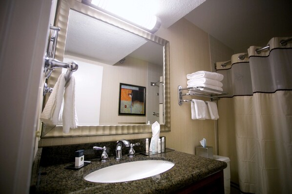 Un baño con lavamanos de encimera de granito, un espejo, toallero con toallas dobladas y cortina de ducha.
