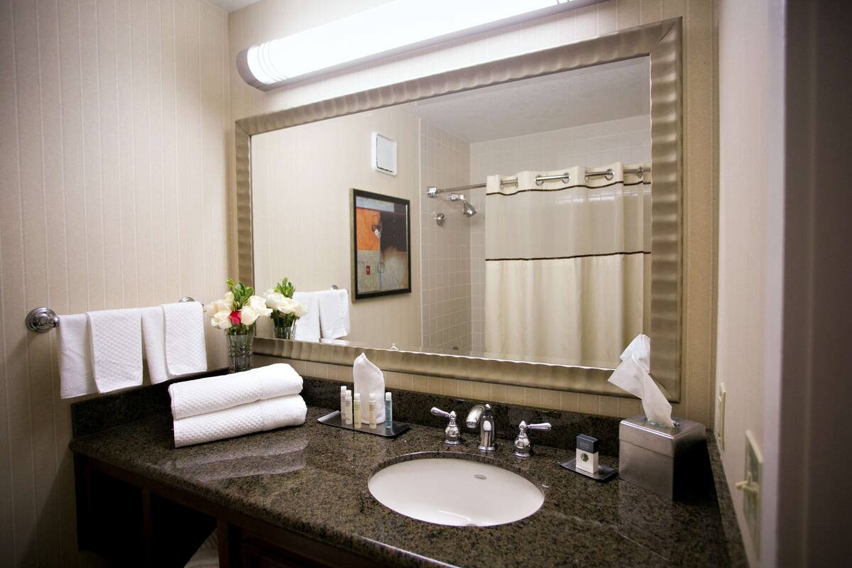 Un baño con un espejo grande, un lavamanos y un perchero con varias toallas.