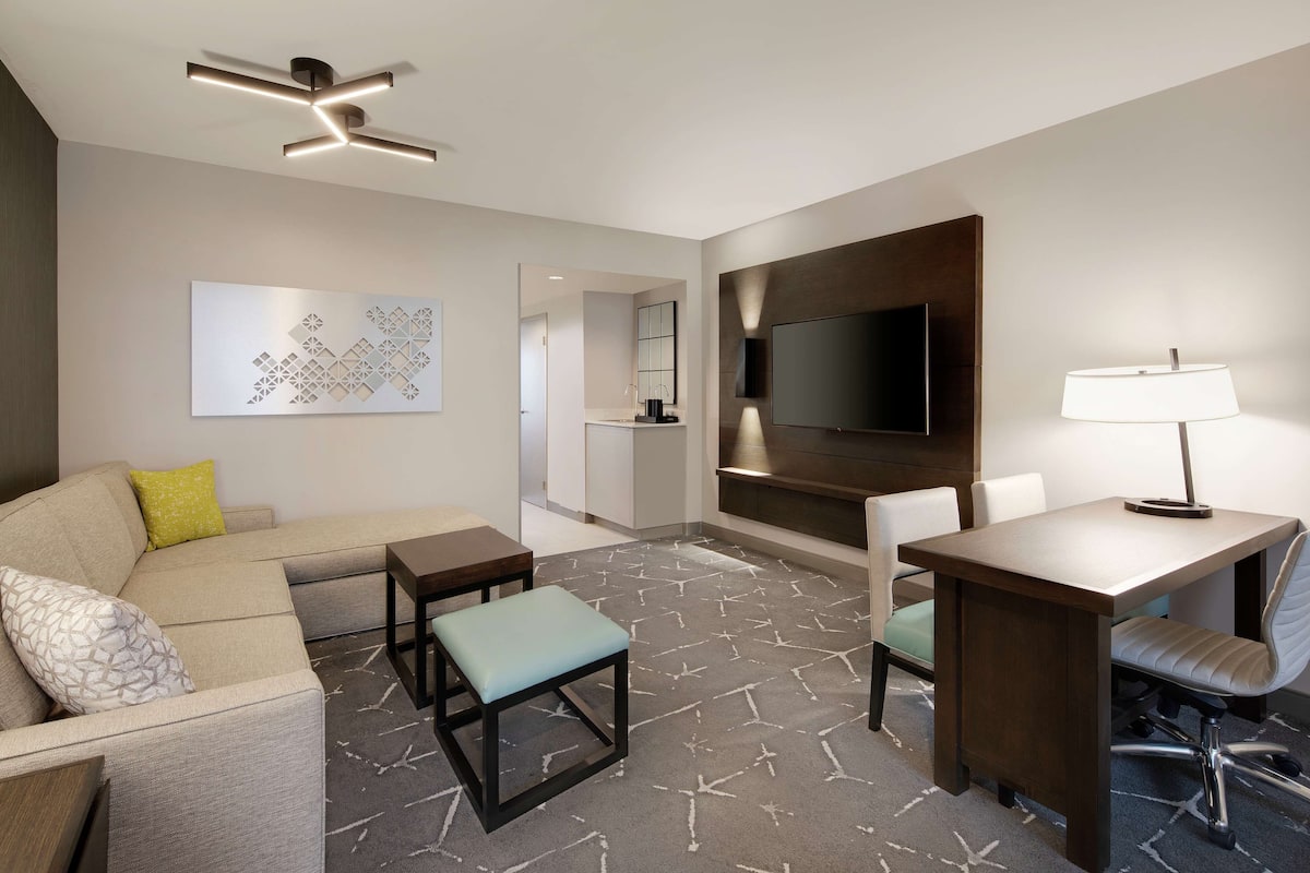 A modern hotel room with a sofa, a desk, a chair, a TV, and a ceiling fan.