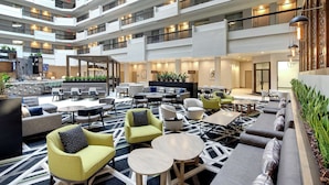 Un hall d’entrée spacieux et intérieur, doté de plusieurs espaces de détente, notamment des canapés, des fauteuils et des tables. On y aperçoit également des plantes et un coin bar.