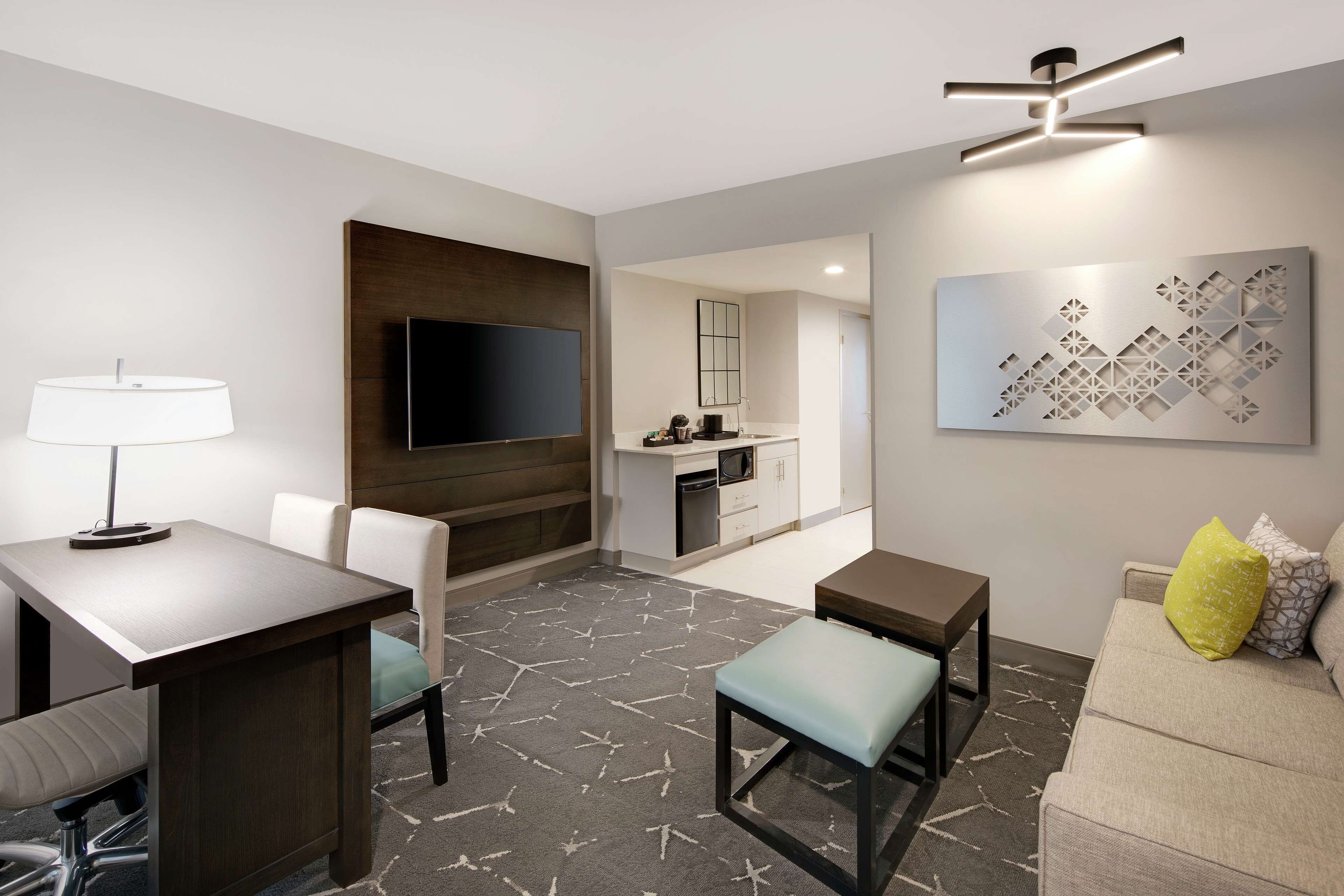 A modern hotel room with a flat-screen TV, a dining area with a table and chairs, a sofa with cushions, a patterned rug, and a ceiling light fixture.