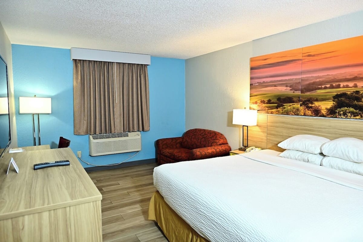Une chambre d’hôtel comprenant un lit, un bureau, une chaise, une fenêtre avec des rideaux, un climatiseur, une lampe et un tableau au mur.