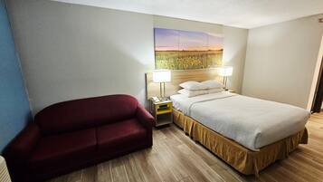 A hotel room with a bed, a red sofa, a nightstand with a lamp, and a painting on the wall.
