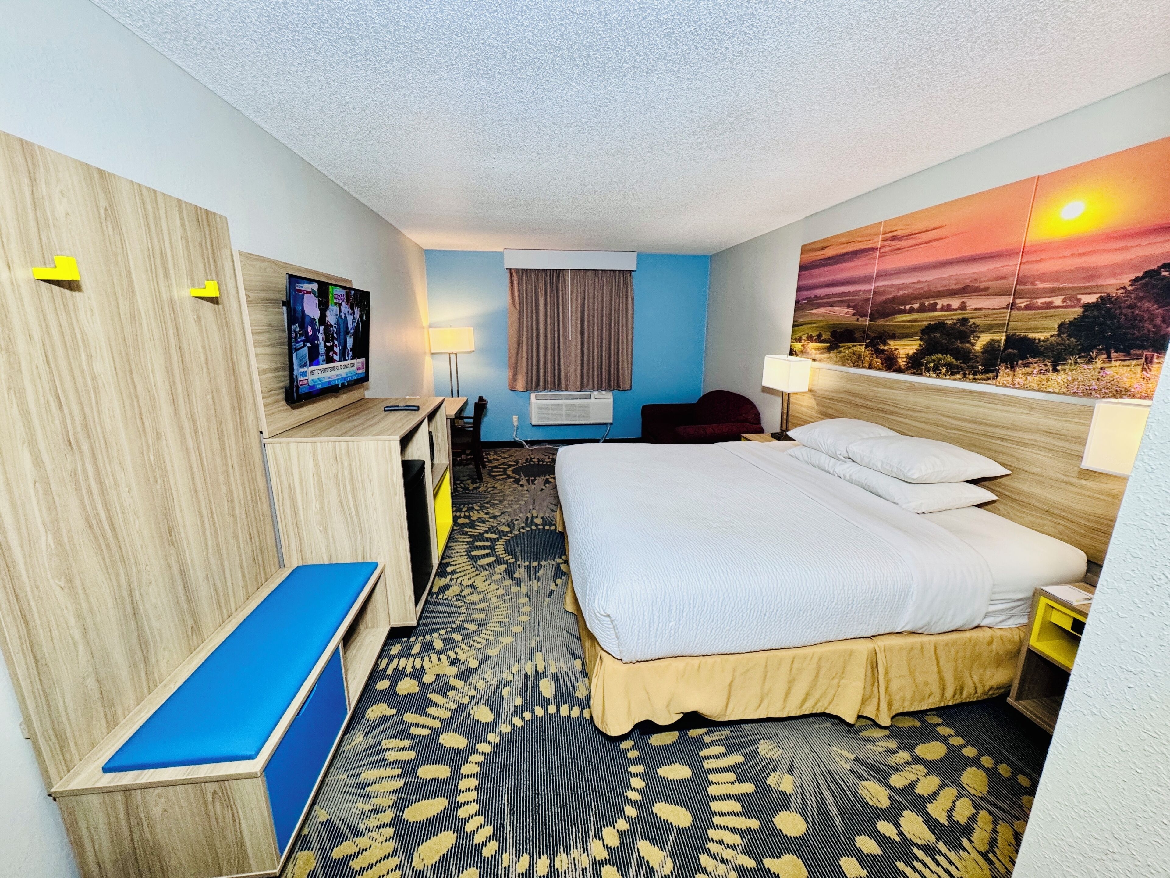 A hotel room with a bed, a desk, a TV, a window with curtains, and a wall painting.