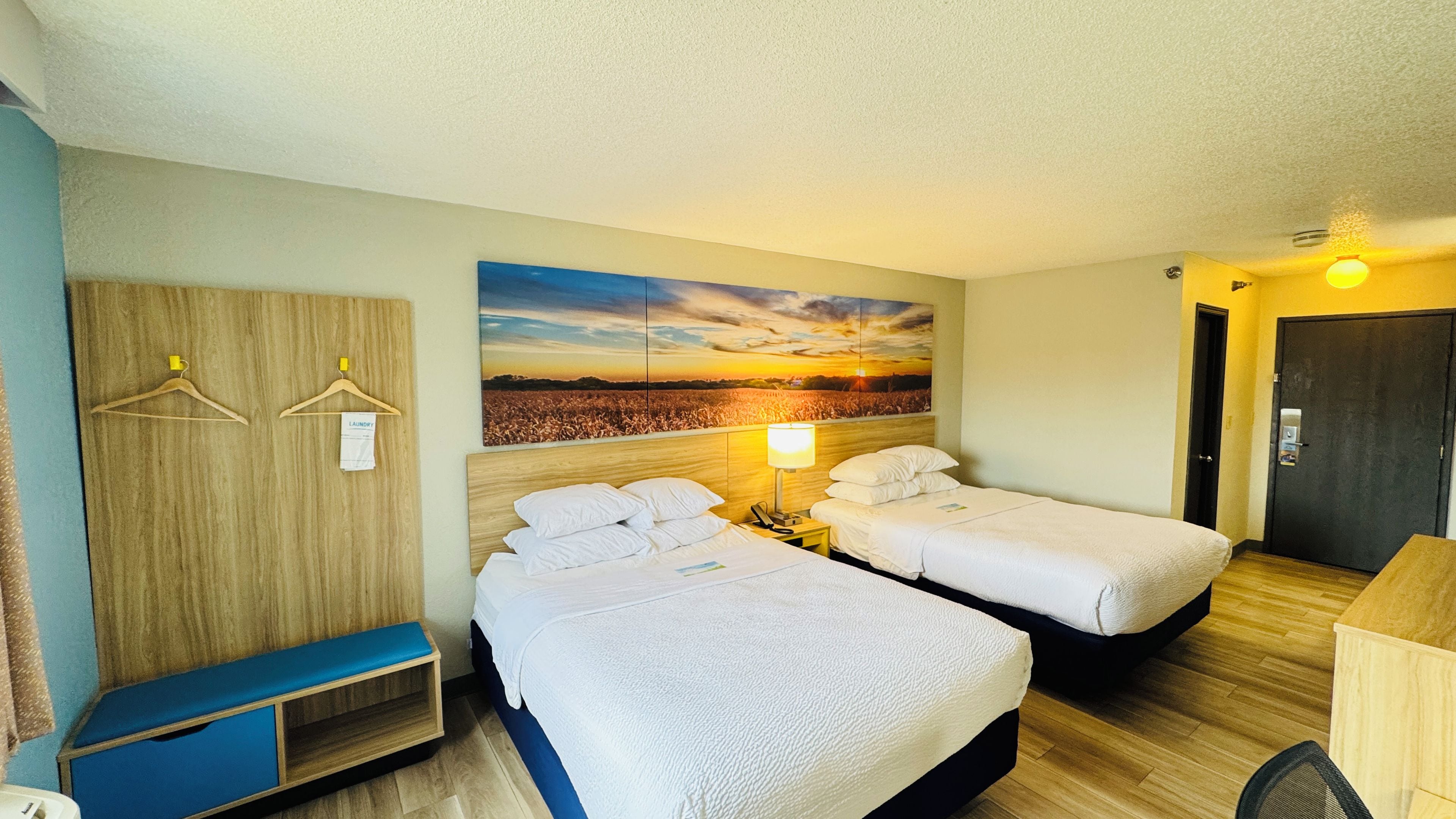 A hotel room with two beds, a bench, a painting of a sunset, a door, and a wall-mounted lamp.