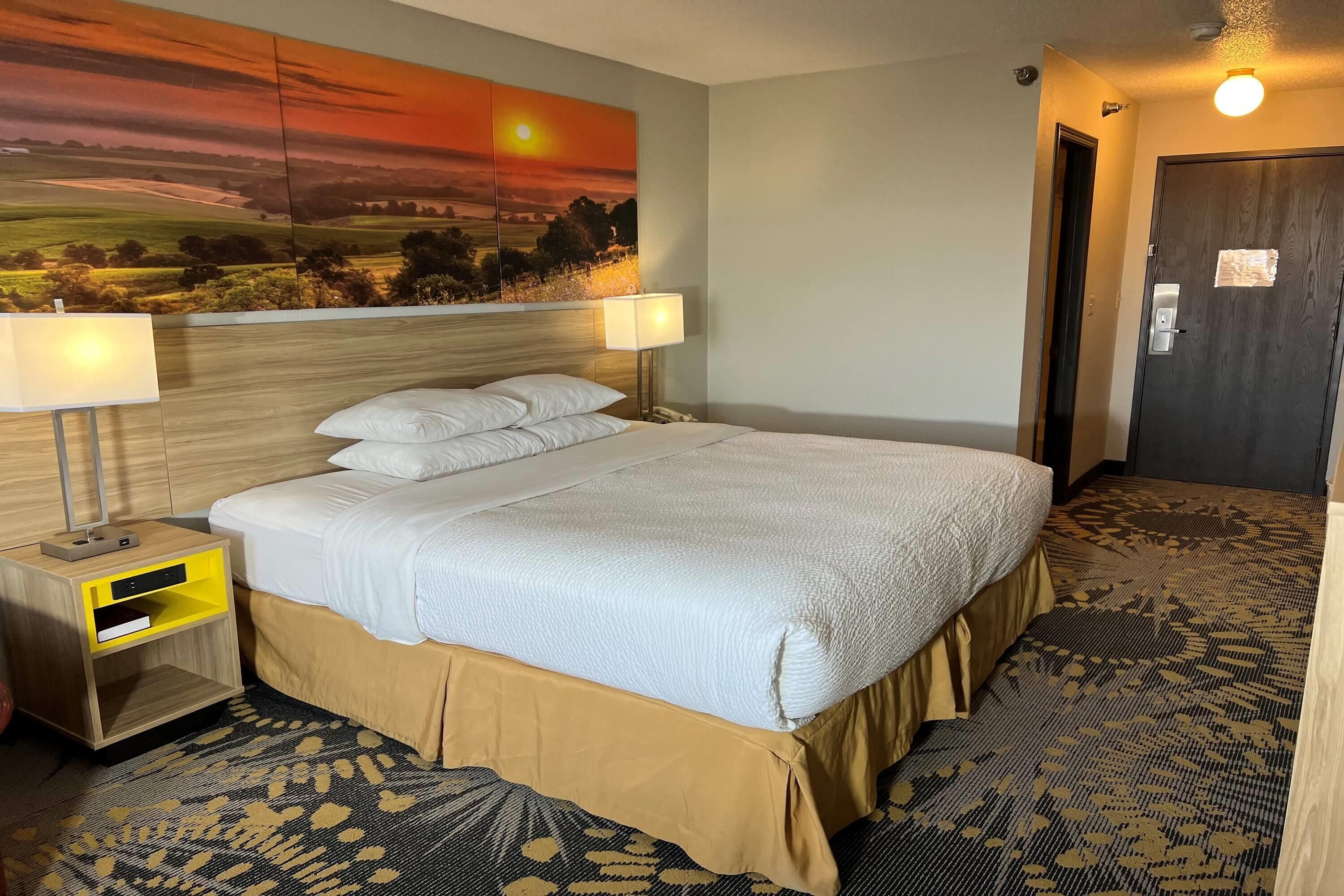 A neatly made bed with white linens, a wooden nightstand with a lamp, and a large landscape painting on the wall.