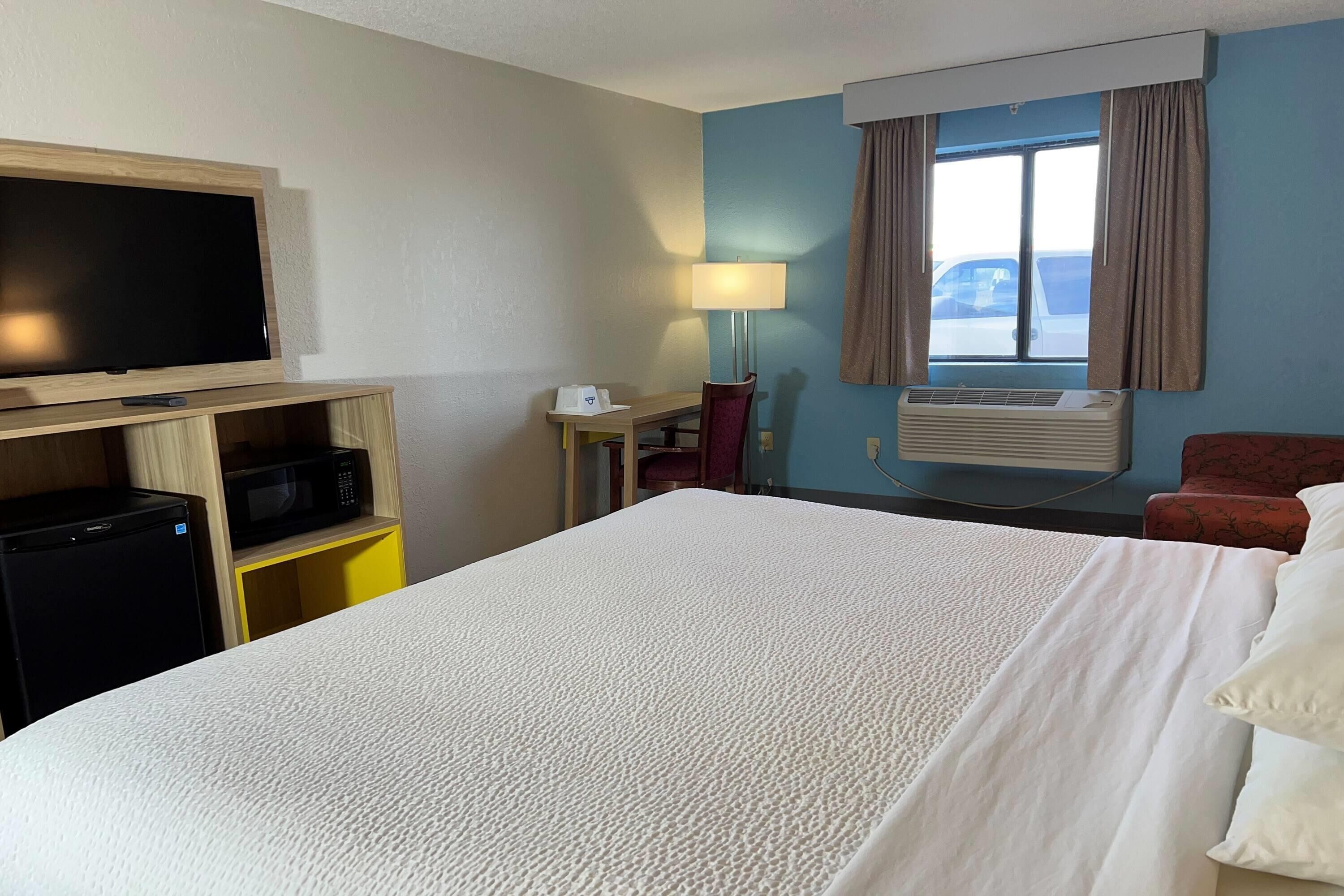 A hotel room with a large bed, a desk with a TV, a microwave, and a small fridge. There is a window with curtains, a lamp, and an air conditioning unit.