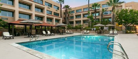 A swimming pool with lounge chairs and umbrellas, surrounded by a building with multiple floors and windows.