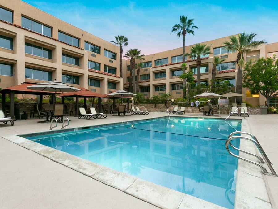 A swimming pool with lounge chairs and umbrellas, surrounded by a building with multiple floors and windows.