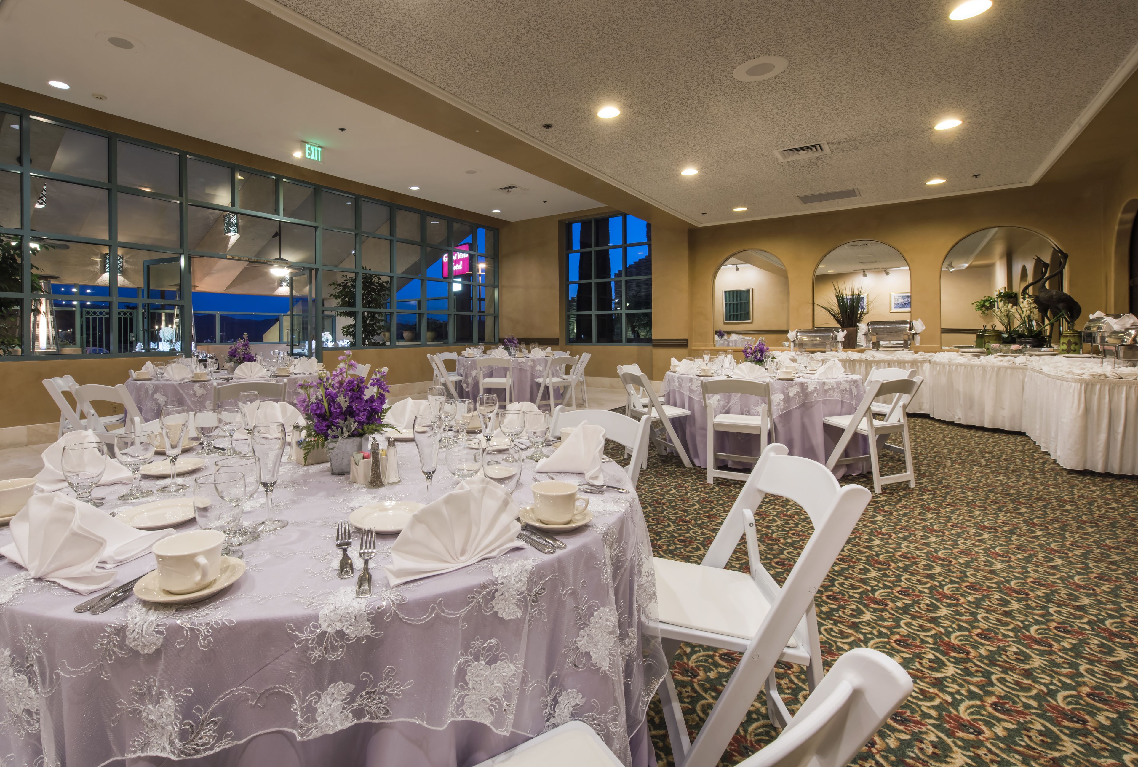 Un salón de banquetes con mesas redondas dispuestas para comer, sillas blancas y ventanas amplias con vistas a la ciudad.