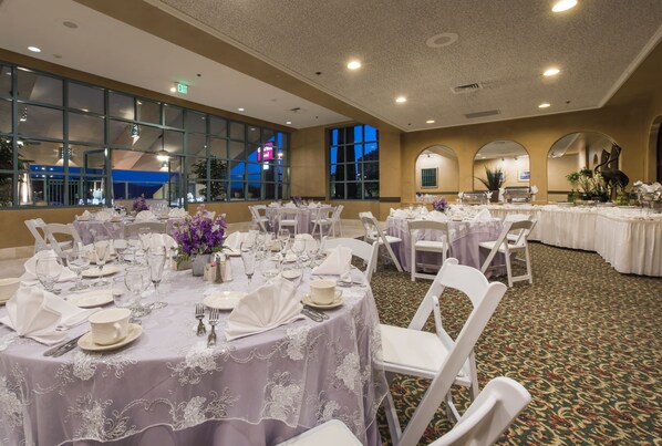 Un salón de banquetes con mesas redondas dispuestas para comer, sillas blancas y ventanas amplias con vistas a la ciudad.