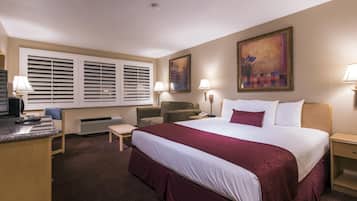 A hotel room with a large bed, two chairs, a desk with a computer, a desk lamp, a telephone, and a painting on the wall.