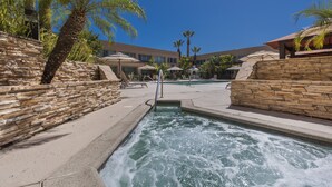 A swimming pool with a stone wall and palm trees, surrounded by lounge chairs and umbrellas. - Grand Vista Hotel (Simi Valley)