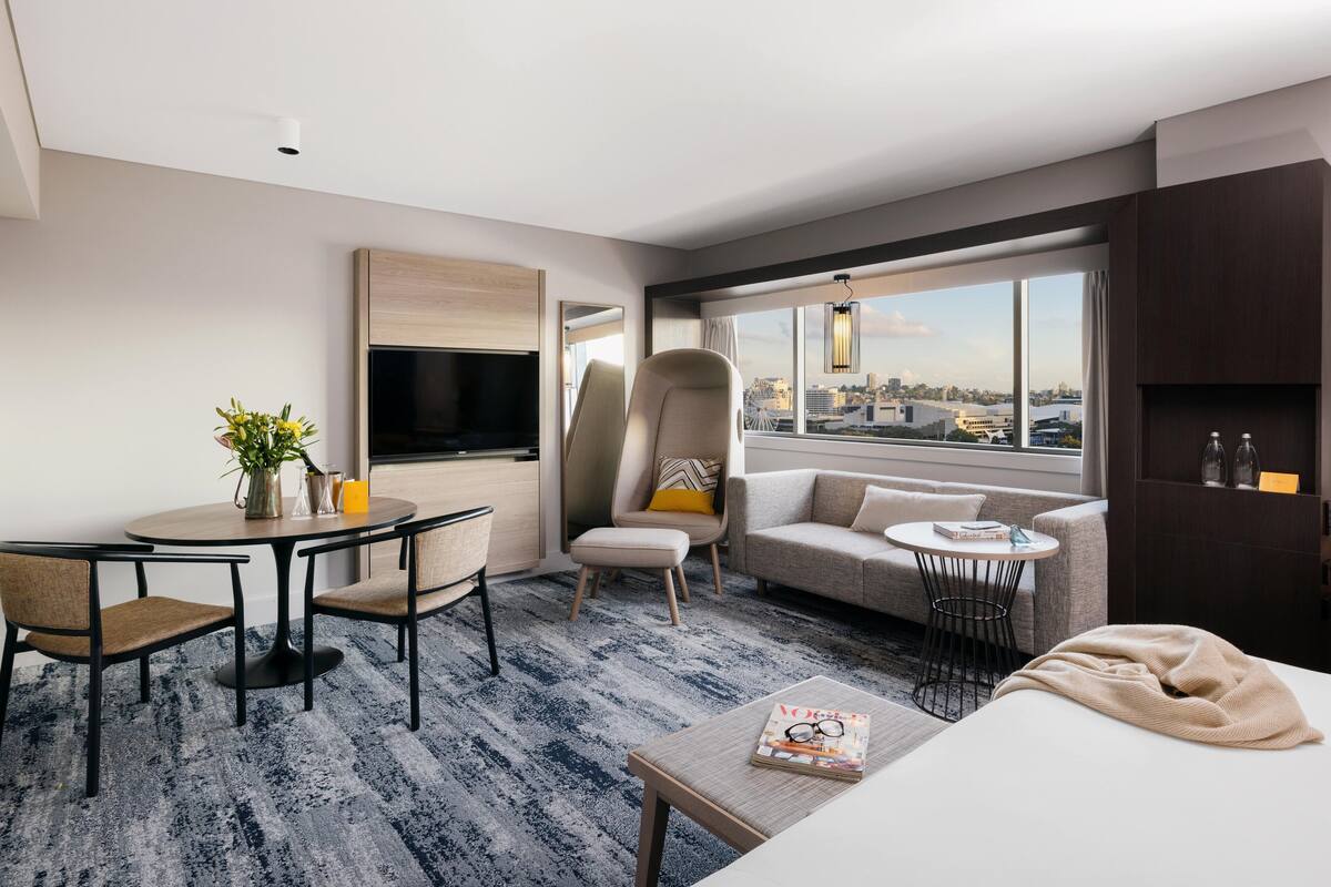A modern hotel room with a dining area, a sofa, a TV, and a large window with a city view.