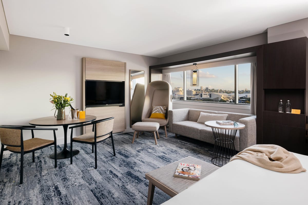 Une chambre d’hôtel moderne dotée d’un coin repas, d’un canapé, d’une télévision et d’une grande fenêtre offrant une vue sur la ville.