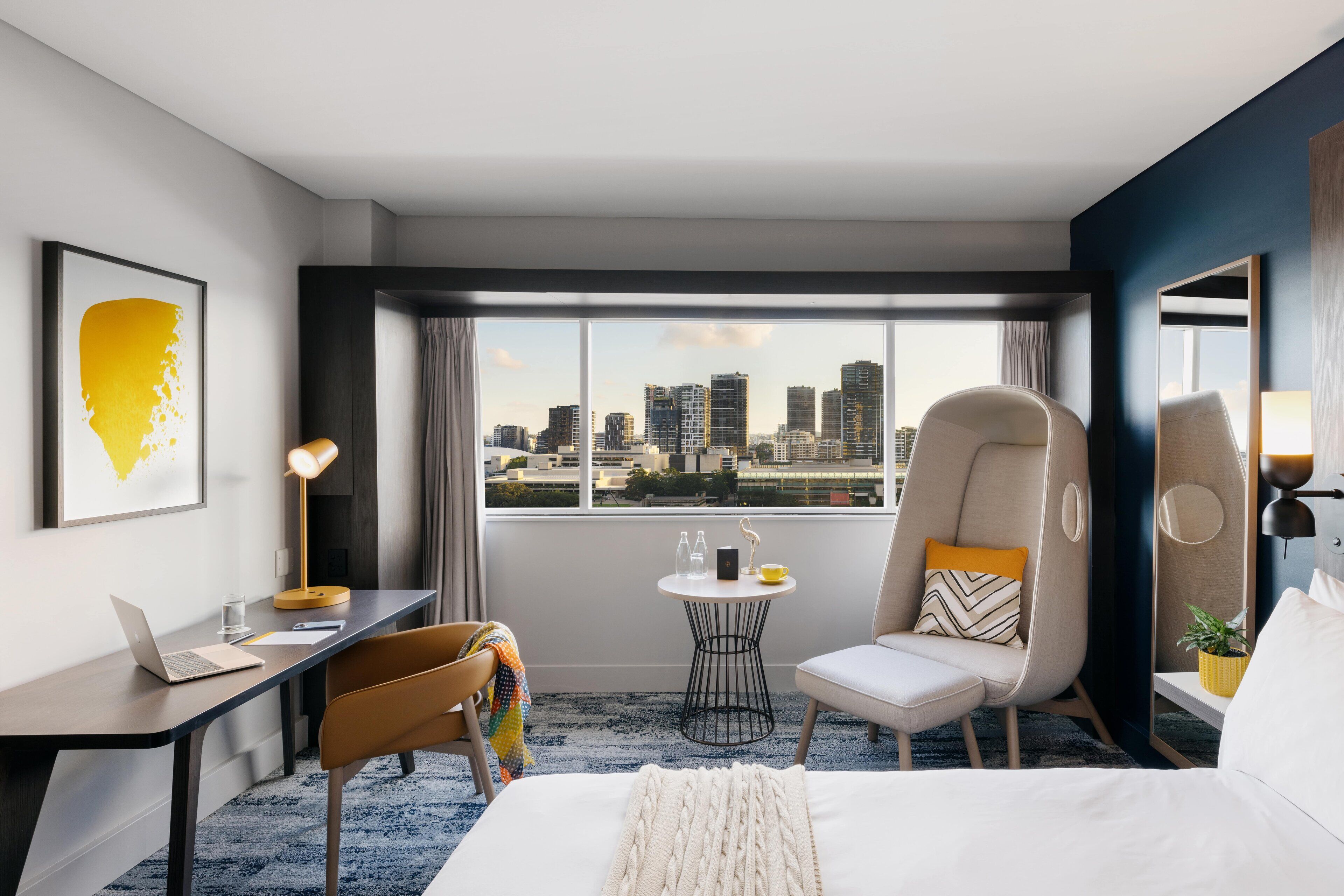 A modern hotel room with a city view, a desk with a laptop, a comfortable chair, a bed, and a small table with a lamp.
