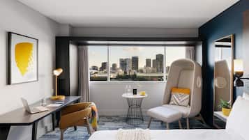 A modern hotel room with a city view, a desk with a laptop, a comfortable chair, a bed, and a small table with a lamp.