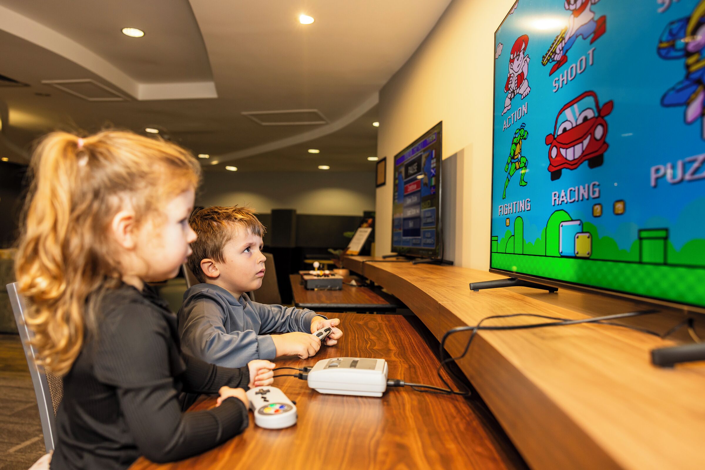 Dos personas jugando un videojuego en una mesa de madera con una pantalla grande mostrando opciones del juego.