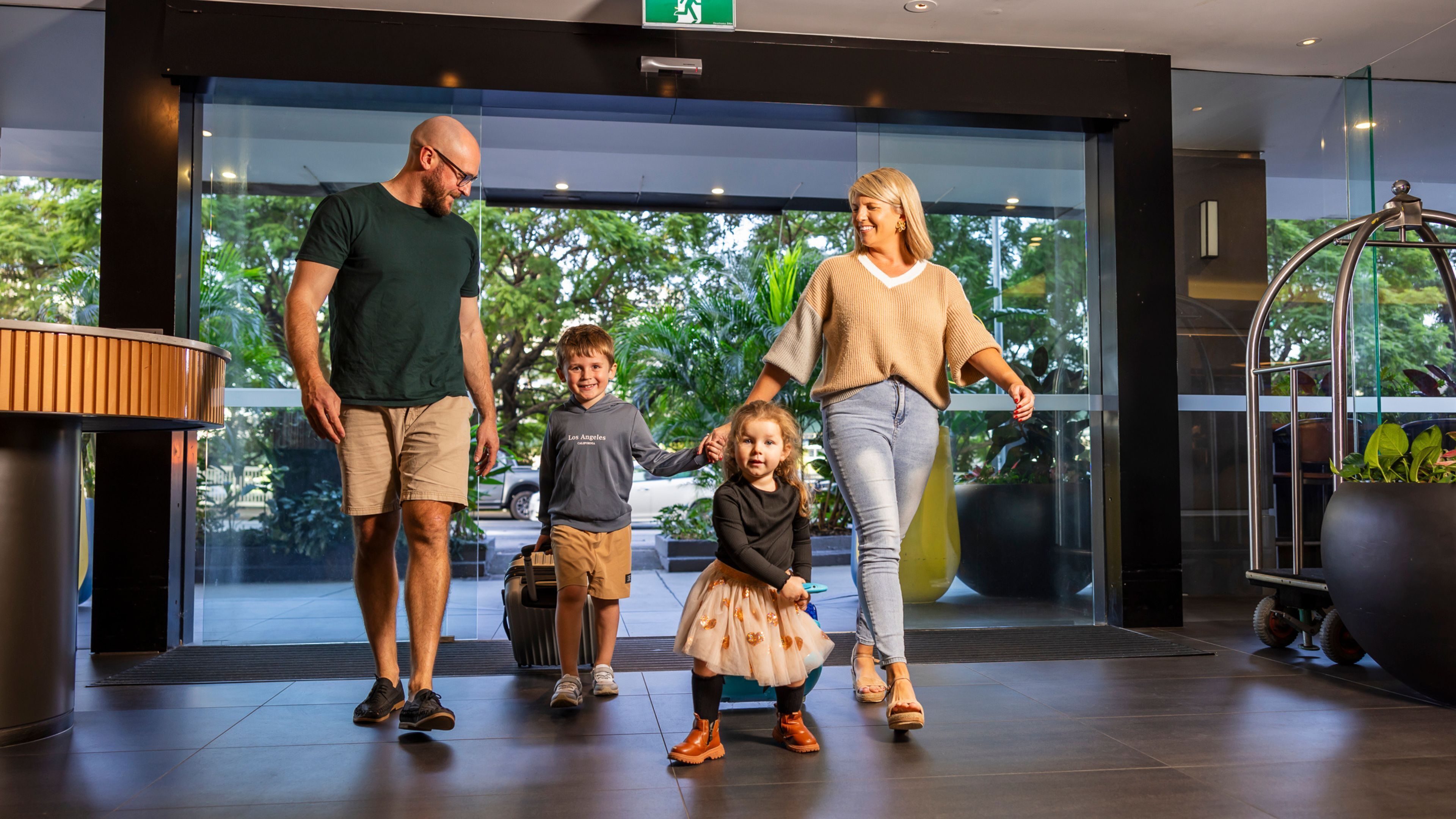 A family of four in a modern lobby with glass doors, a visible exit sign, and a large potted plant.