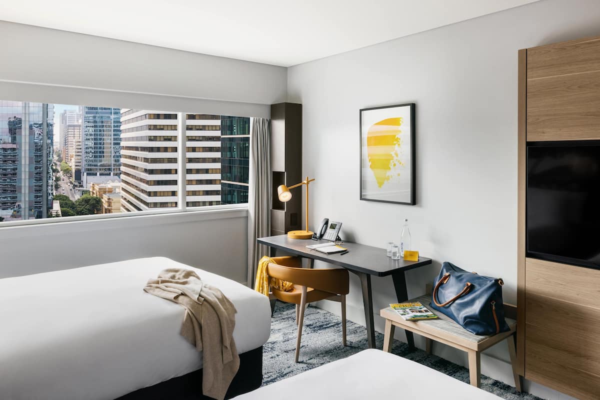 A hotel room with two beds, a desk, a chair, a TV, a desk lamp, a handbag, and a window with a city view.