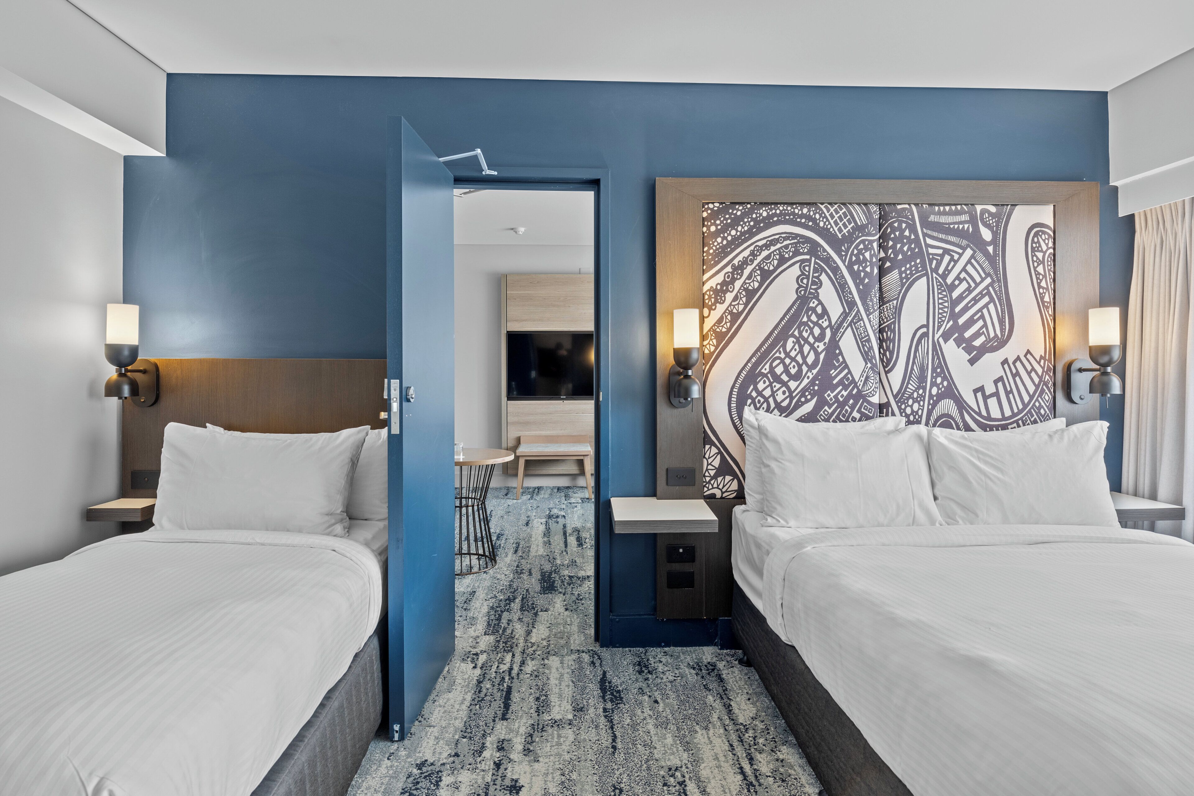 Une chambre d’hôtel avec deux lits, un mur d’accent bleu, une tête de lit à motifs et un petit bureau avec une lampe.