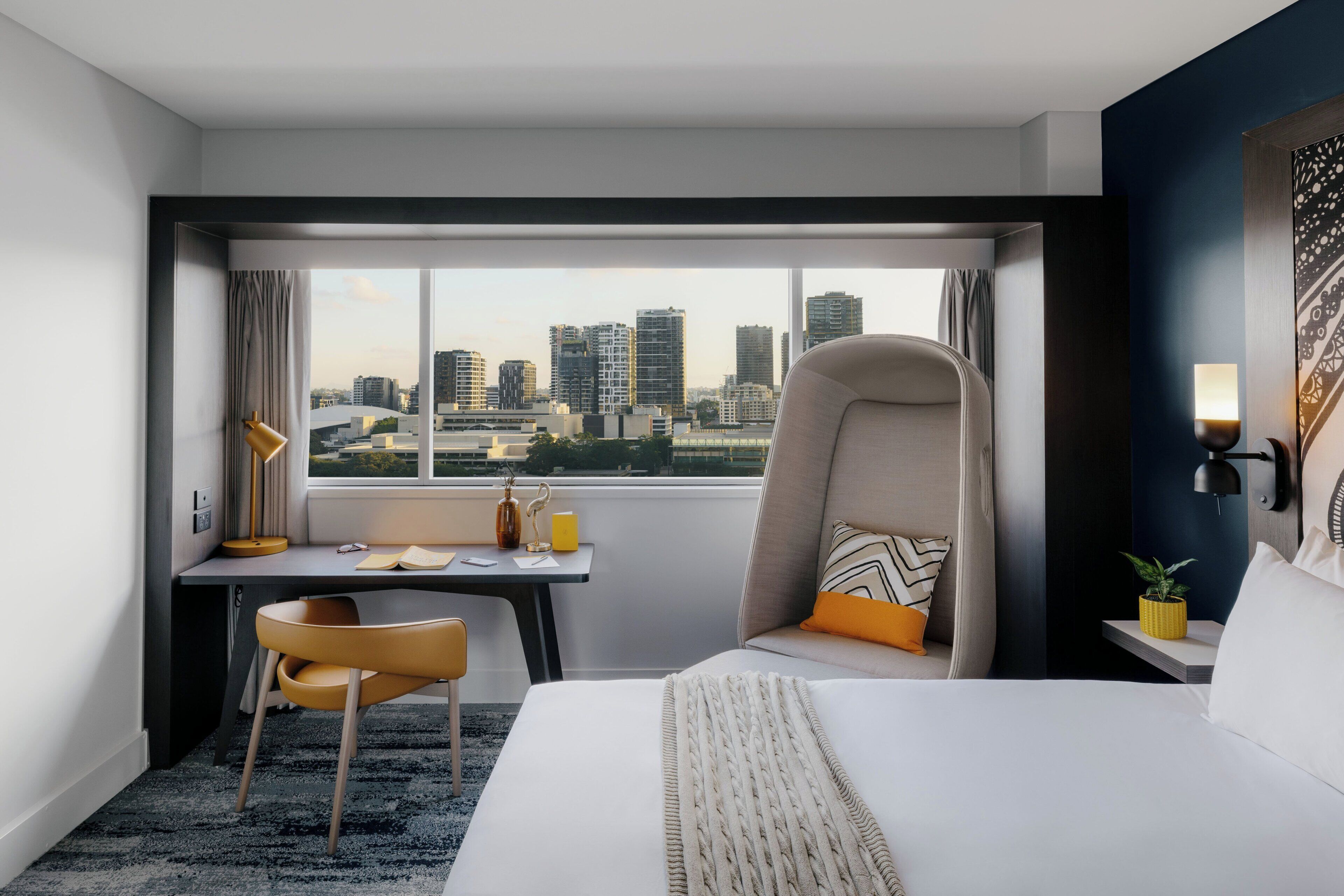 Une chambre d’hôtel avec un lit, un bureau, une chaise, une fenêtre donnant sur la ville et une lampe.