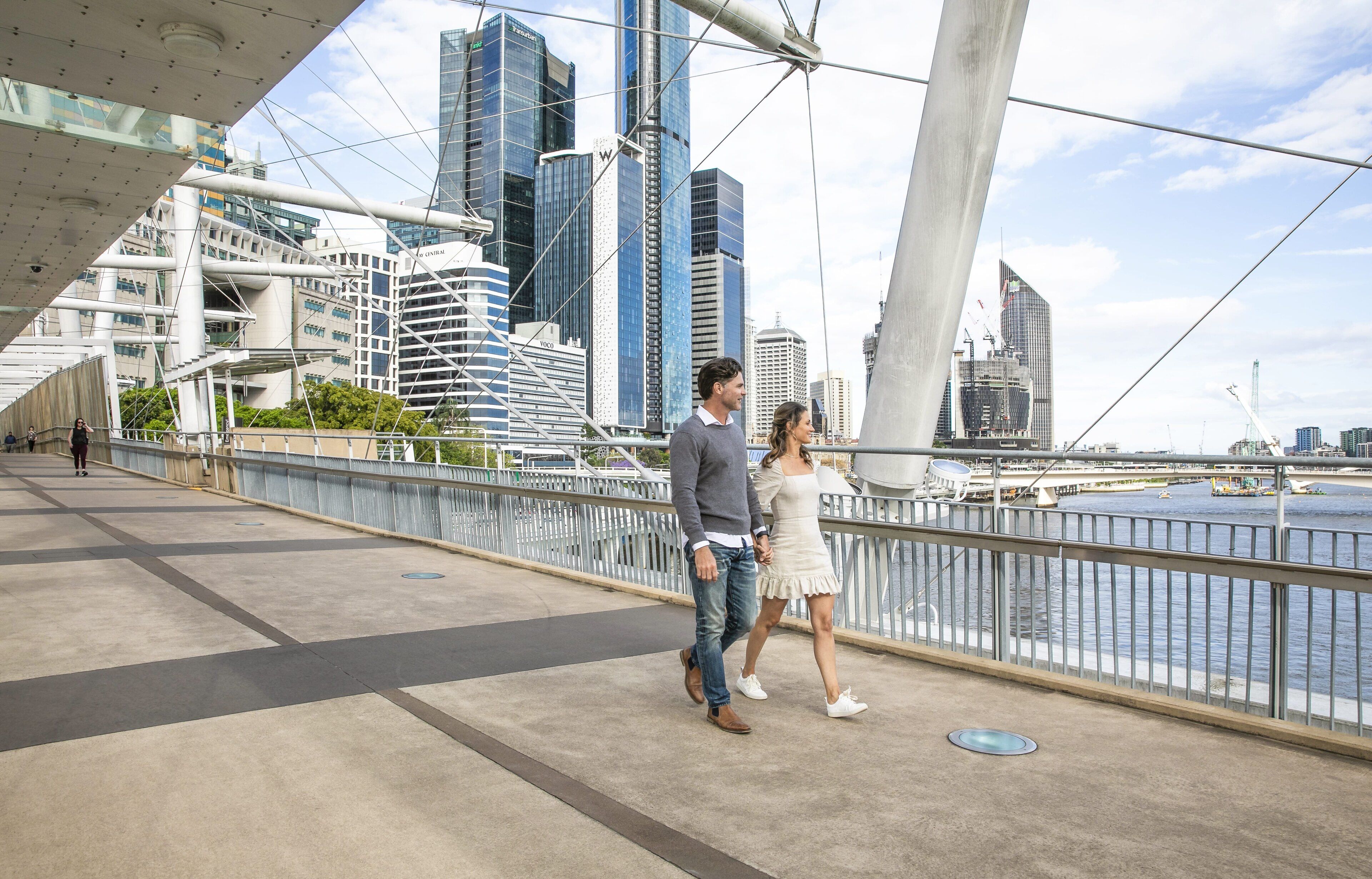 A couple walking on a bridge with a cityscape in the background.