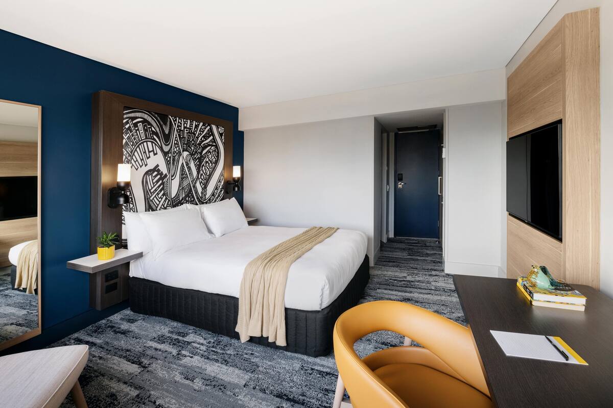 A modern hotel room with a large bed, a desk, a chair, a mirror, and a wall art piece.