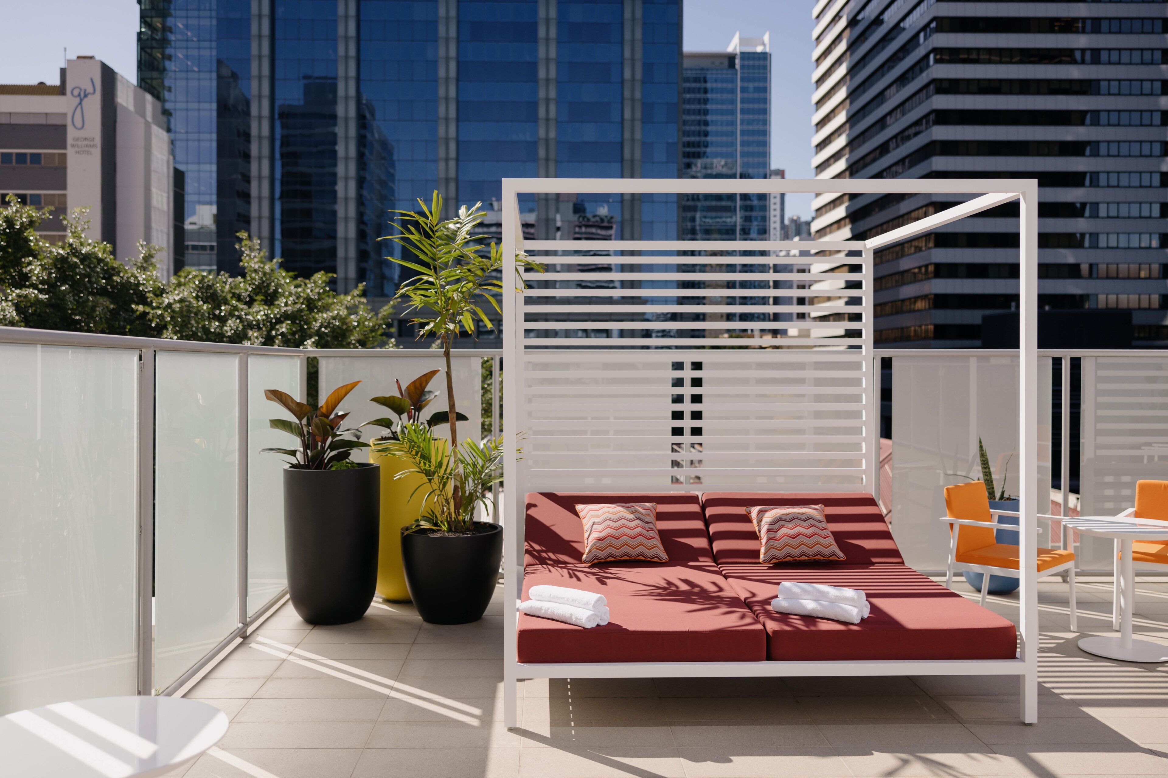A rooftop terrace with a red cushioned seating area, white chairs, and potted plants.