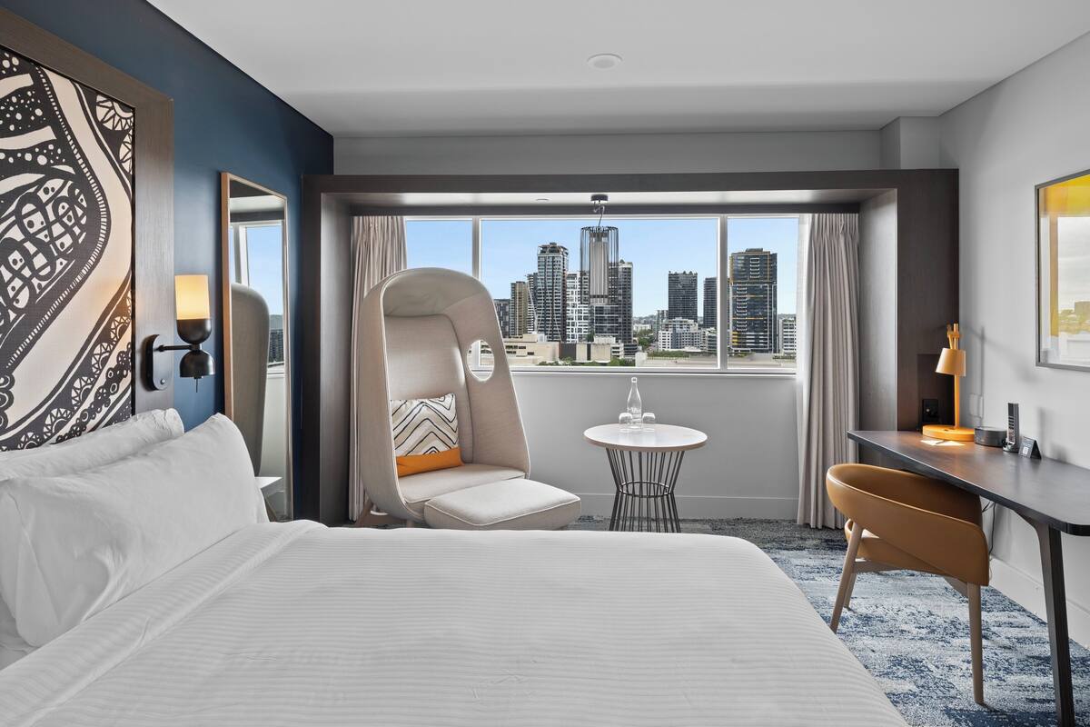 A hotel room with a large bed, a chair, a small table, and a view of the city skyline.