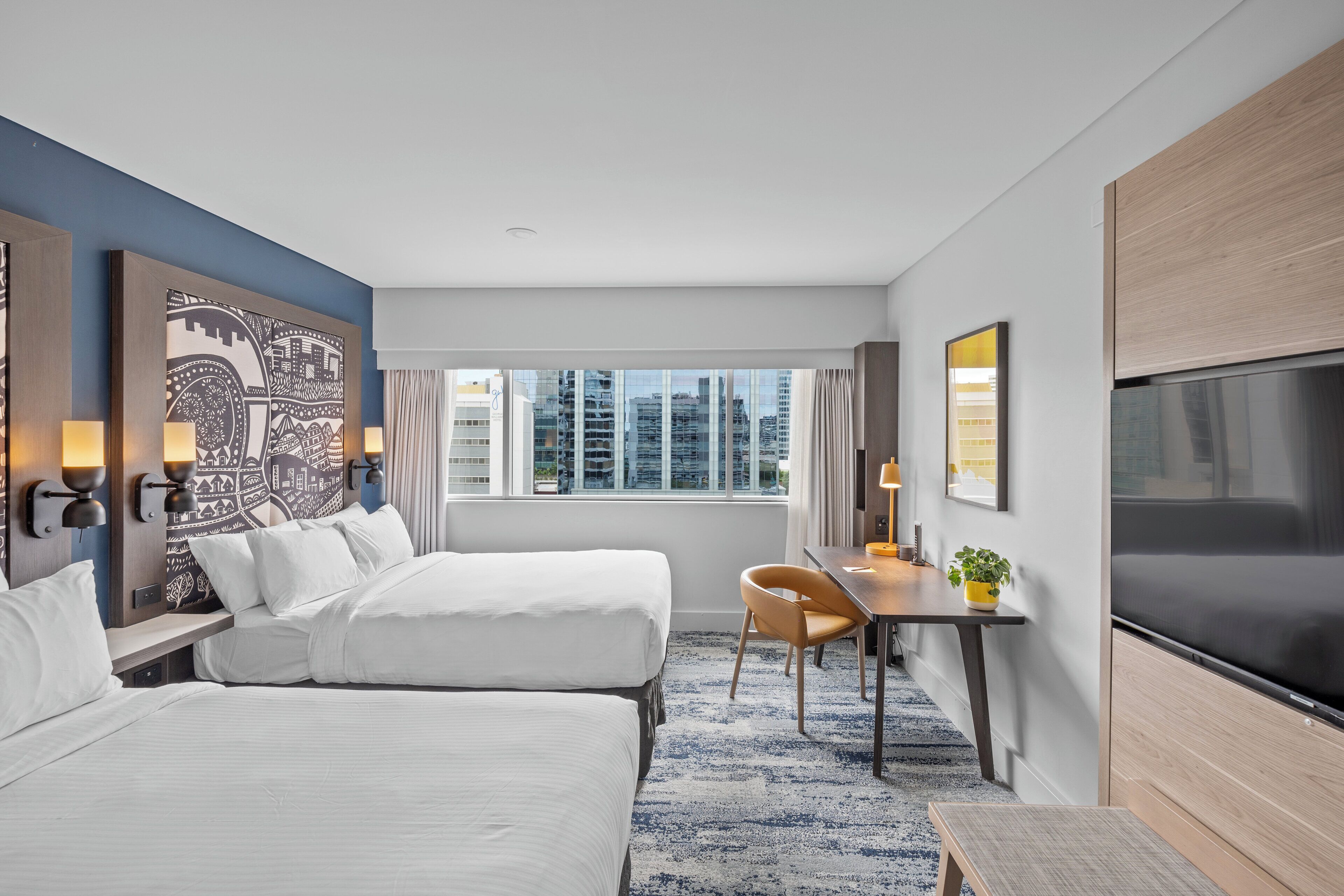 A hotel room with two beds, a desk, a chair, a TV, and a large window offering a city view.