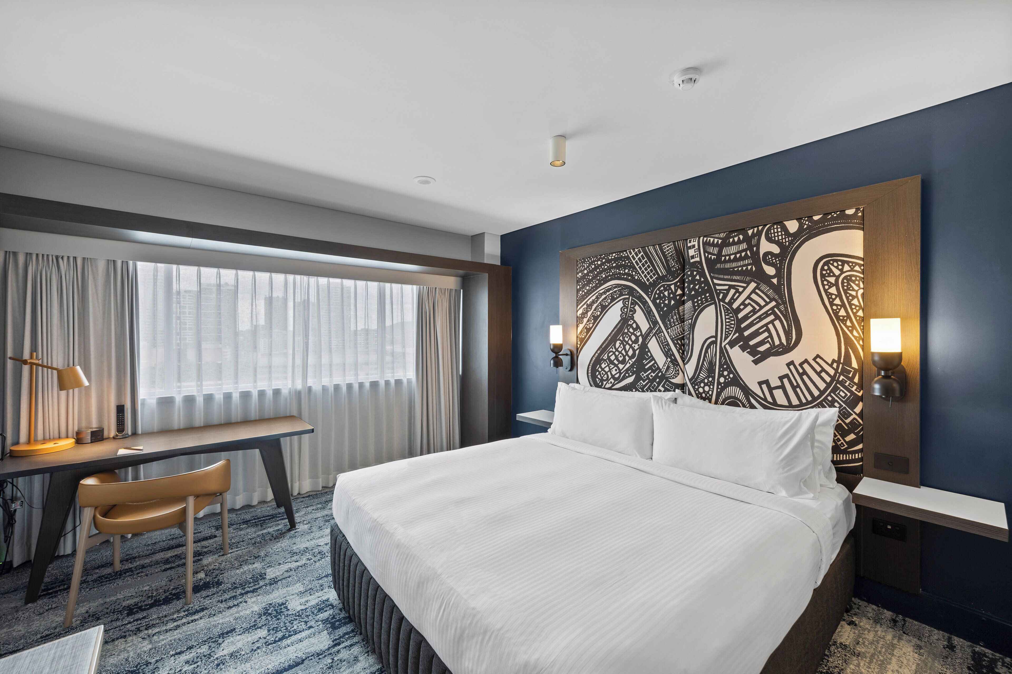 Une chambre d’hôtel avec un grand lit, un bureau avec une chaise, une grande fenêtre avec des rideaux et un élément décoratif mural.