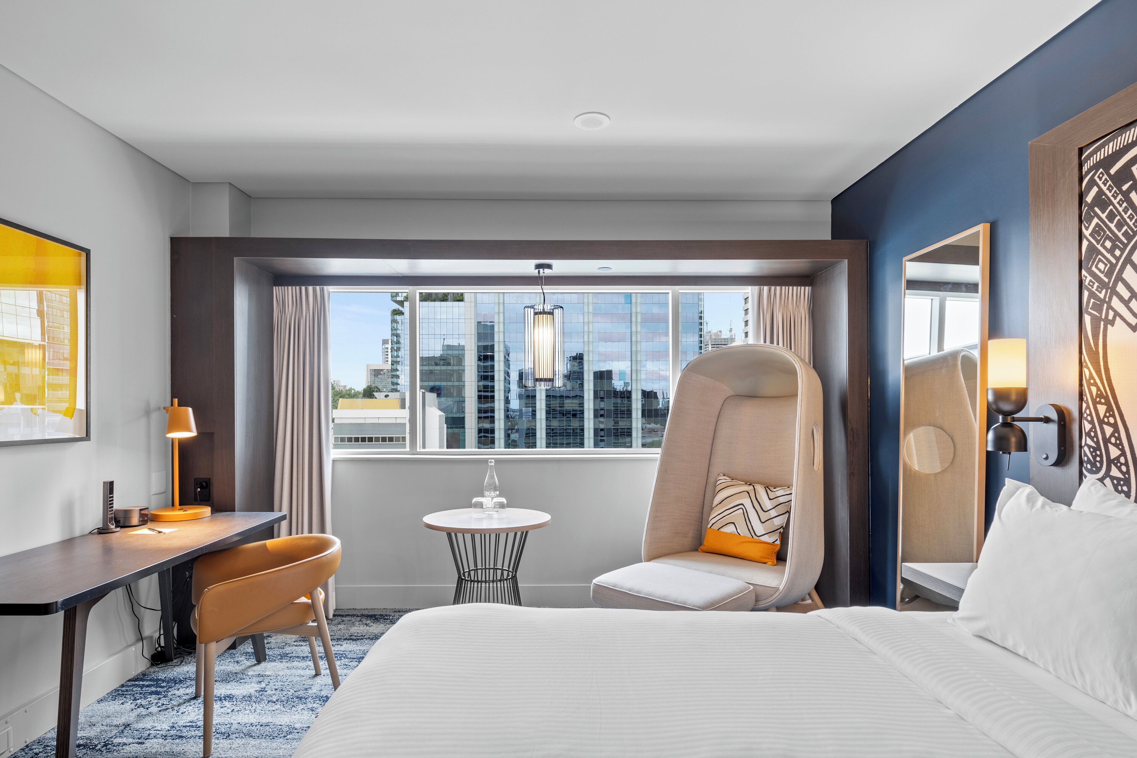 Une chambre d’hôtel comprenant un lit, un bureau, une chaise, une petite table et une grande fenêtre donnant sur la ville.