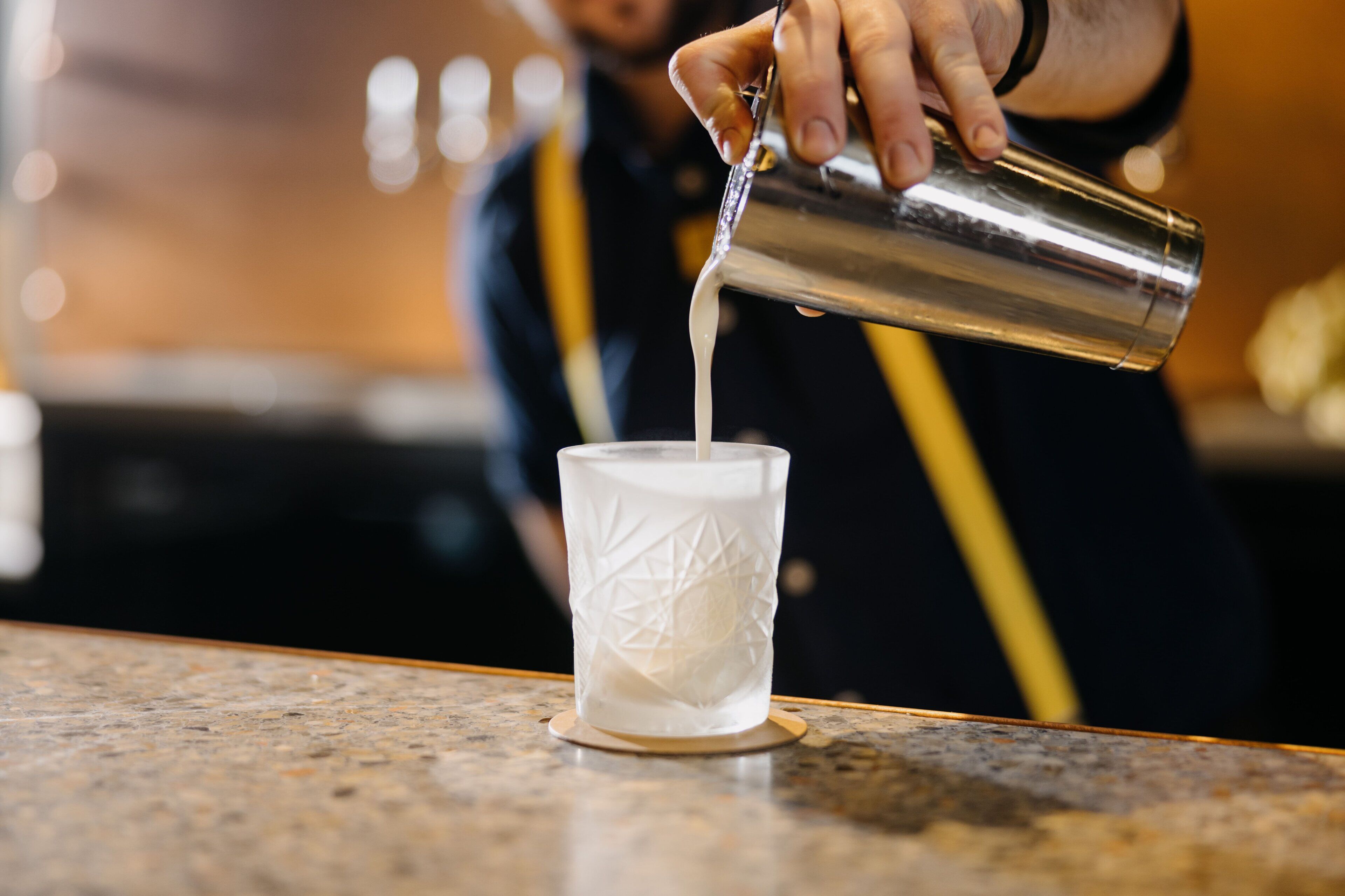 A person pouring a liquid from a metal container into a glass on a countertop.