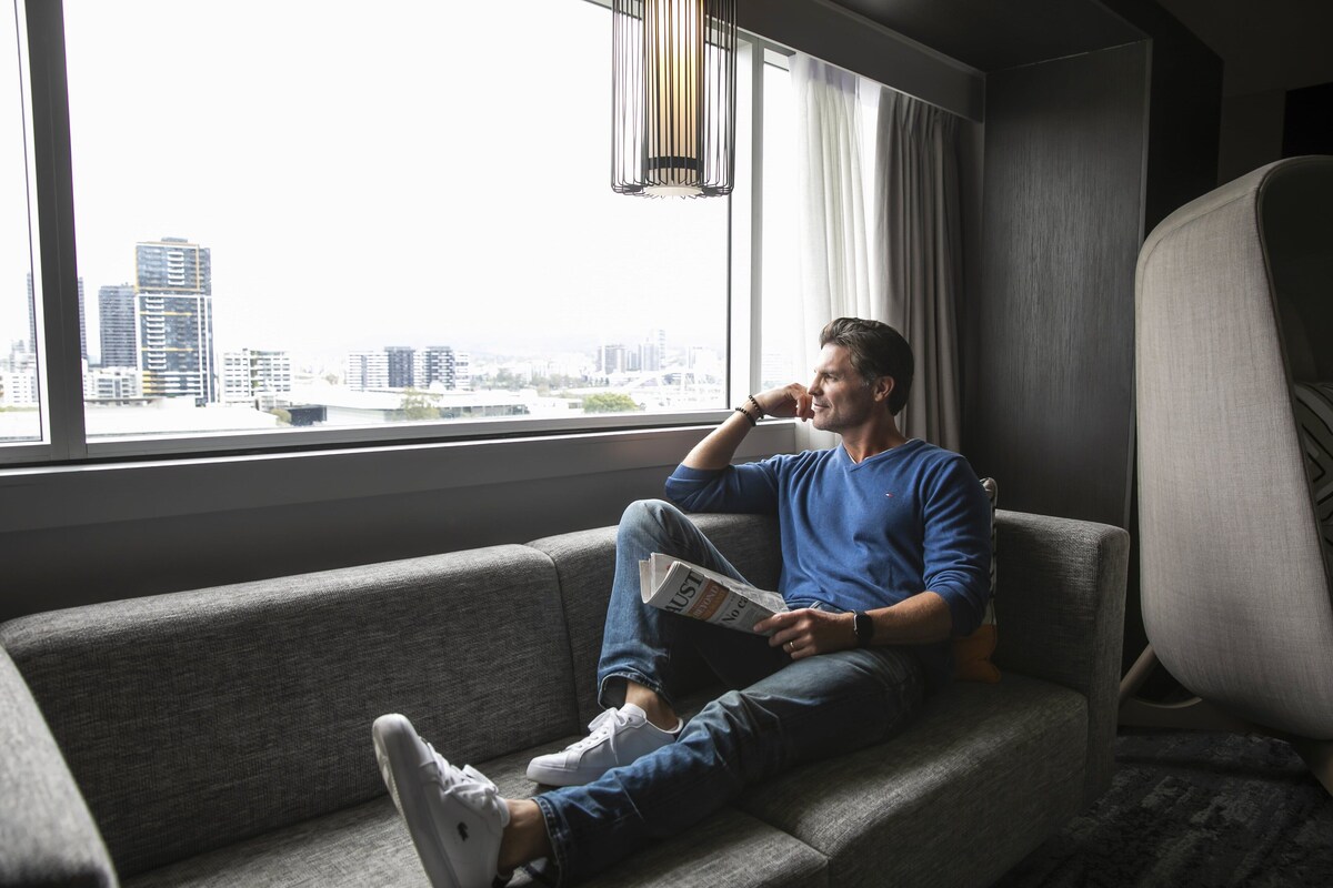 A person sitting on a grey sofa in a room with a large window, reading a newspaper.