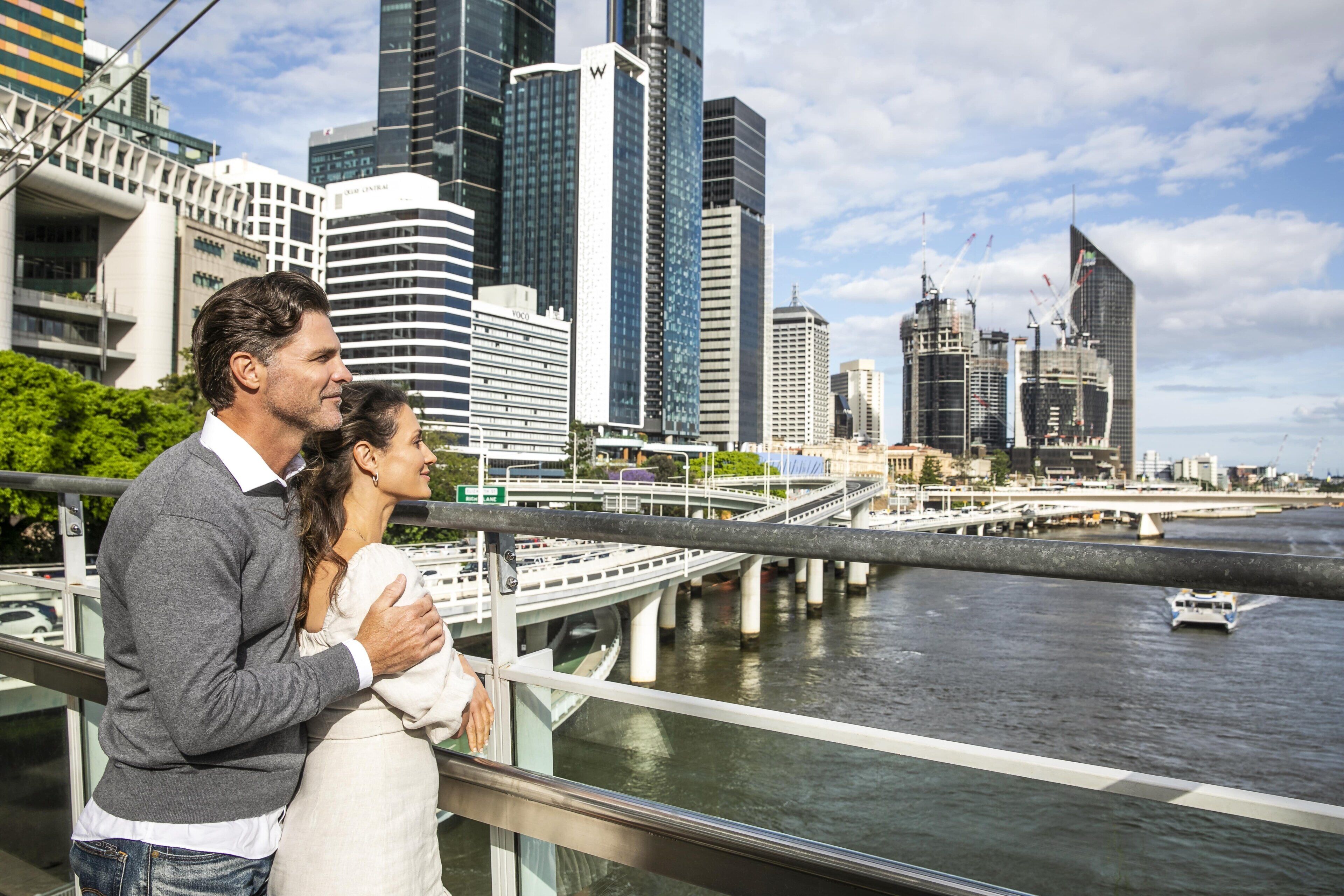 Una pareja de pie sobre un puente con vistas a un paisaje urbano con edificios modernos y un río.