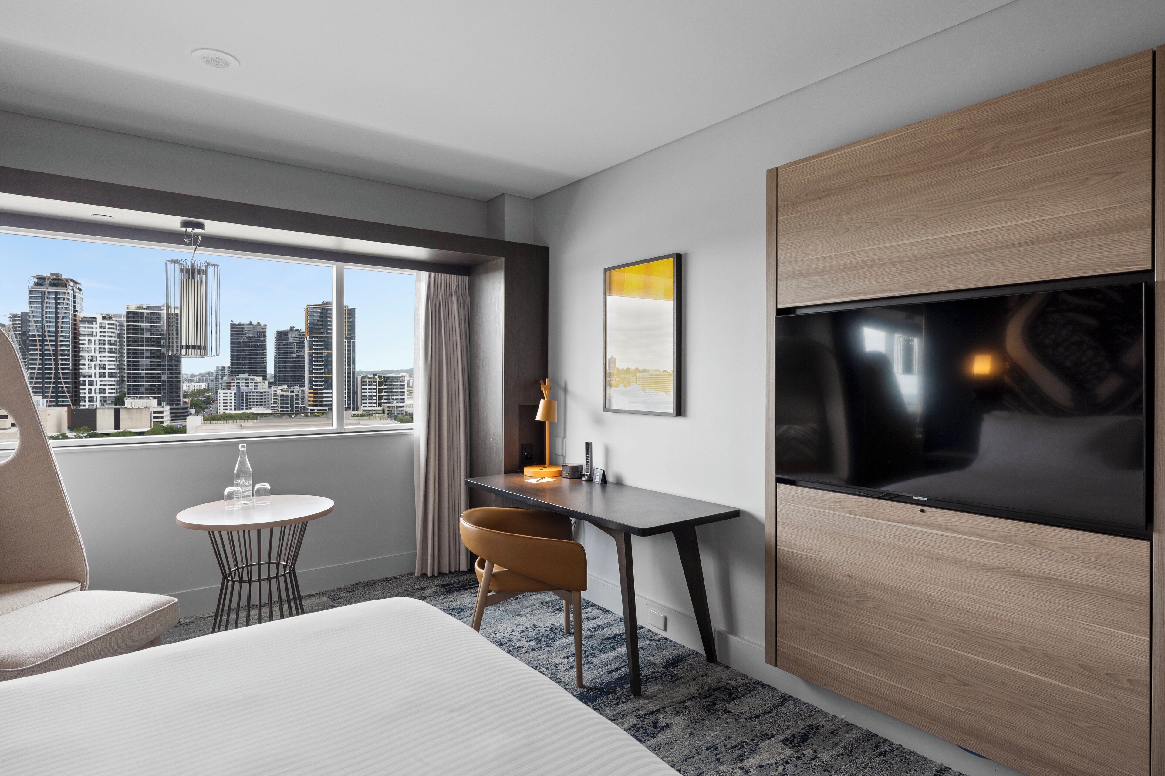 A modern hotel room with a large bed, a desk with a chair, a TV, and a view of the city skyline.