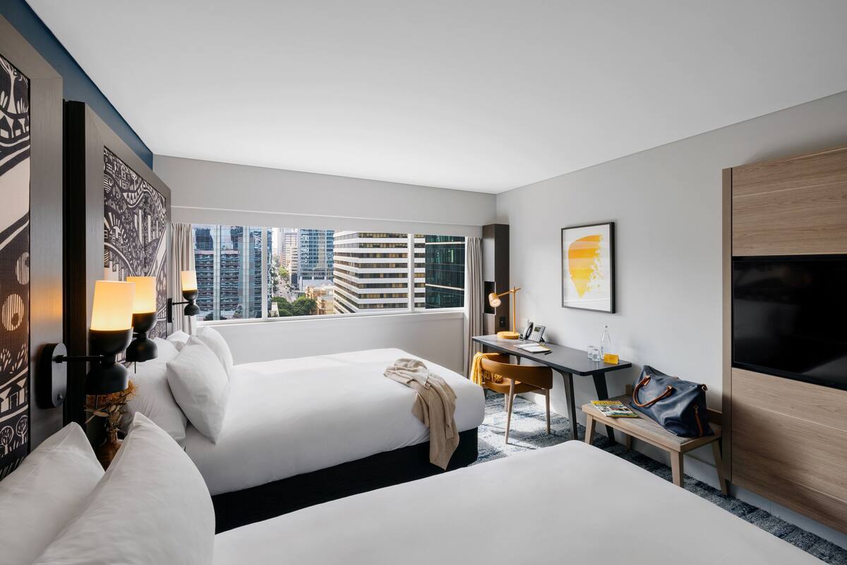 A hotel room with two beds, a desk, a chair, a TV, and a large window with a city view.