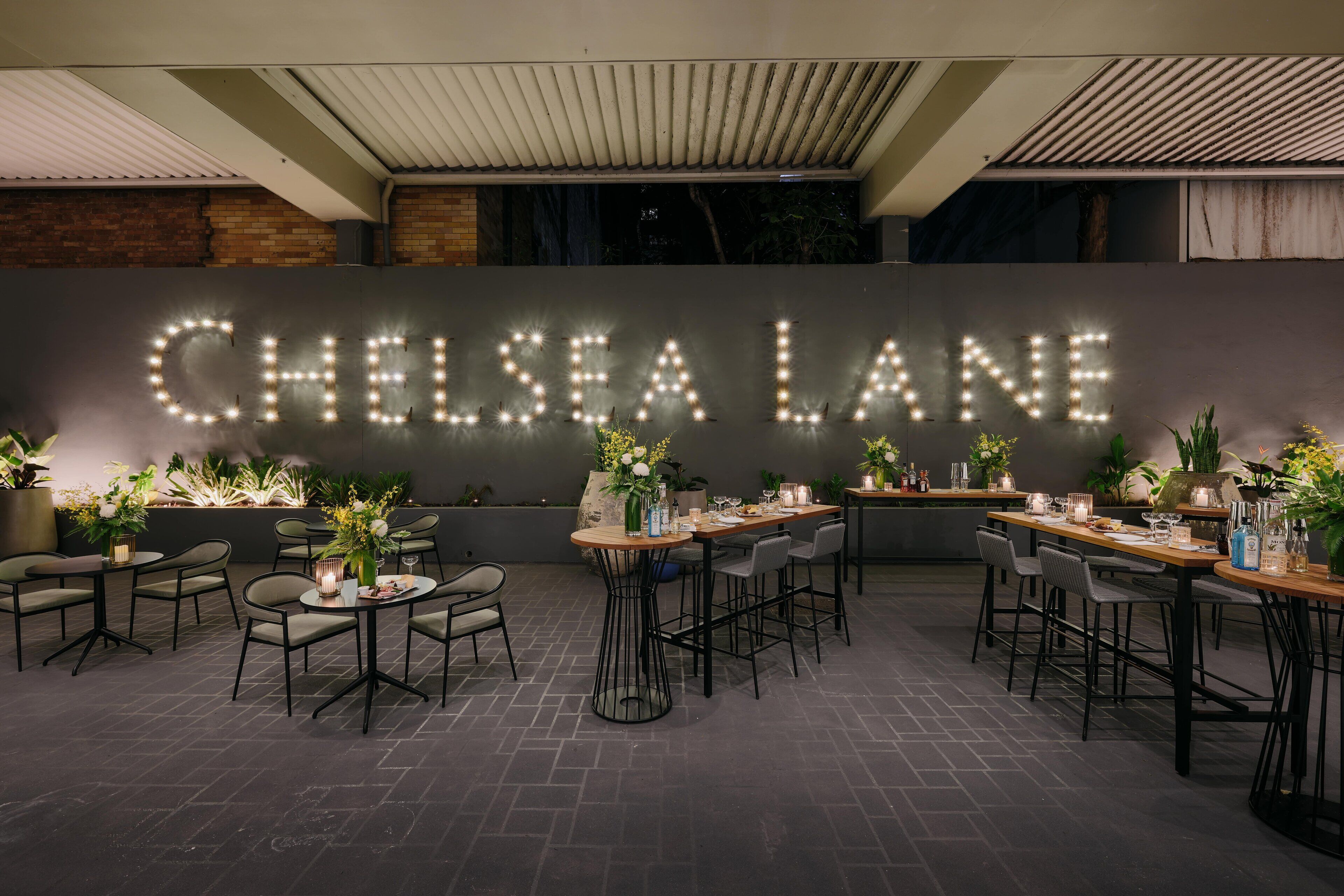 Un espace extérieur aménagé pour les repas, avec des tables et des chaises, une grande enseigne lumineuse indiquant « CHELSEA LANE », et des plantes en pot.
