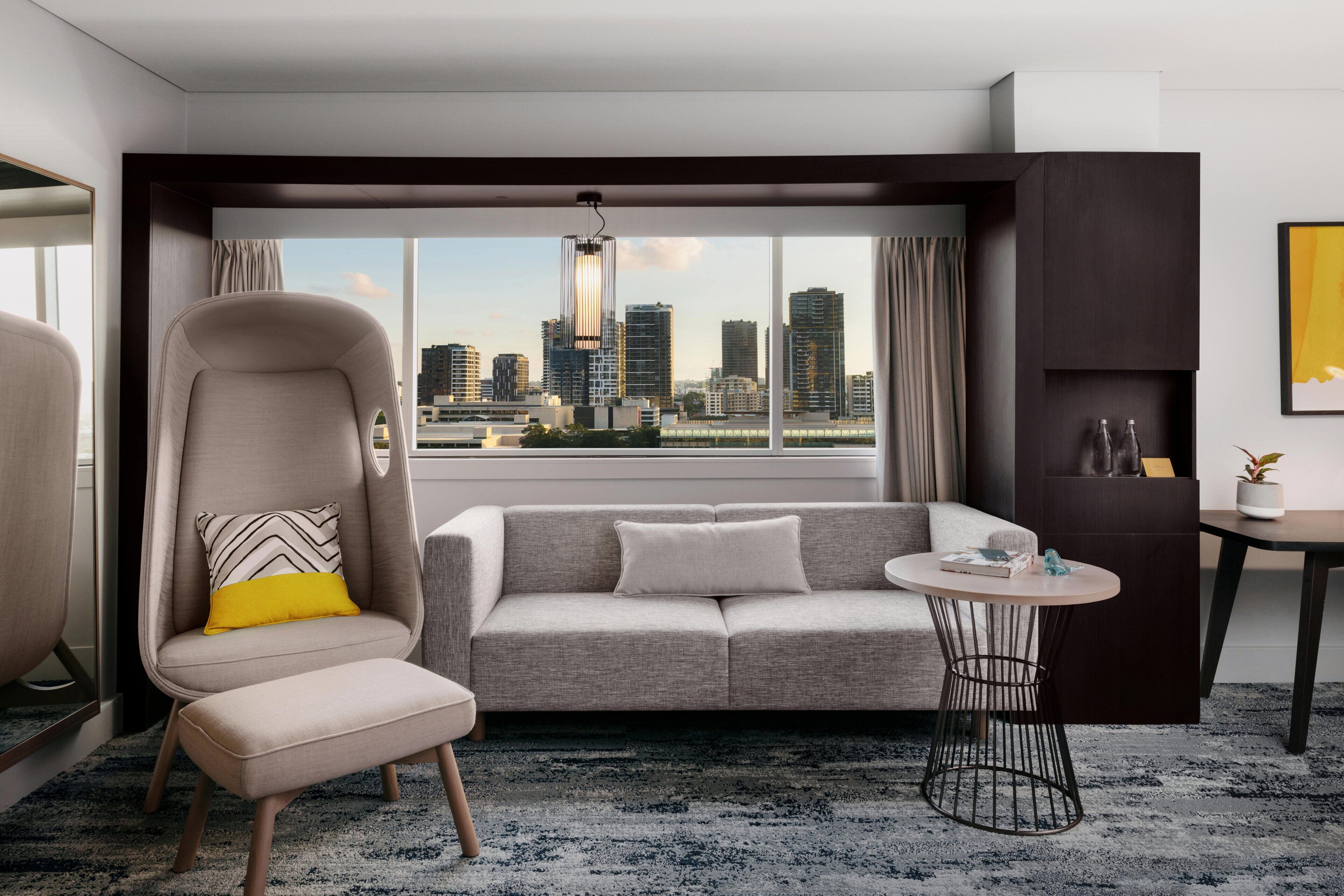 A modern hotel room with a city view, a grey sofa, a high-back chair, a small round table, and a large window.