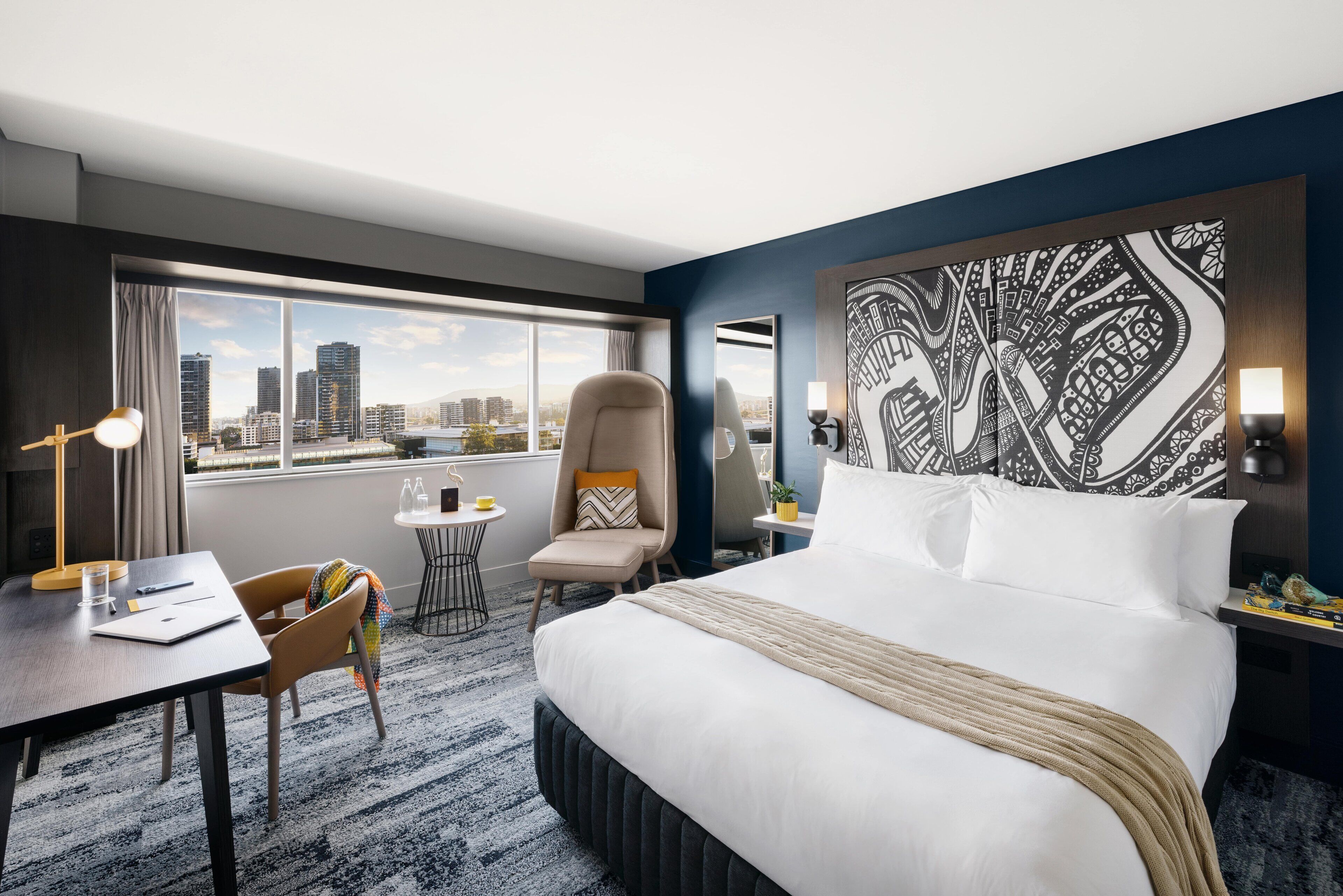 A hotel room with a large bed, a desk, a chair, a small table, and a view of the city skyline.