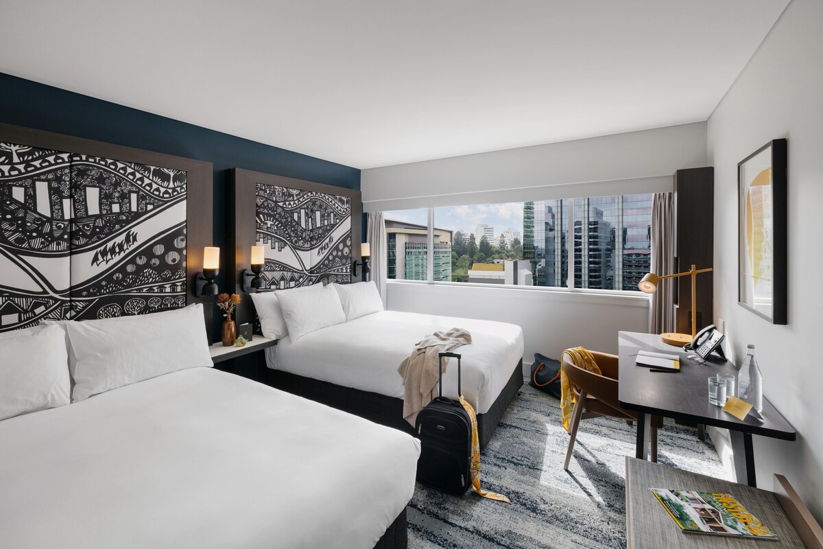 A hotel room with two beds, a desk, a chair, a suitcase, and a view of the city.